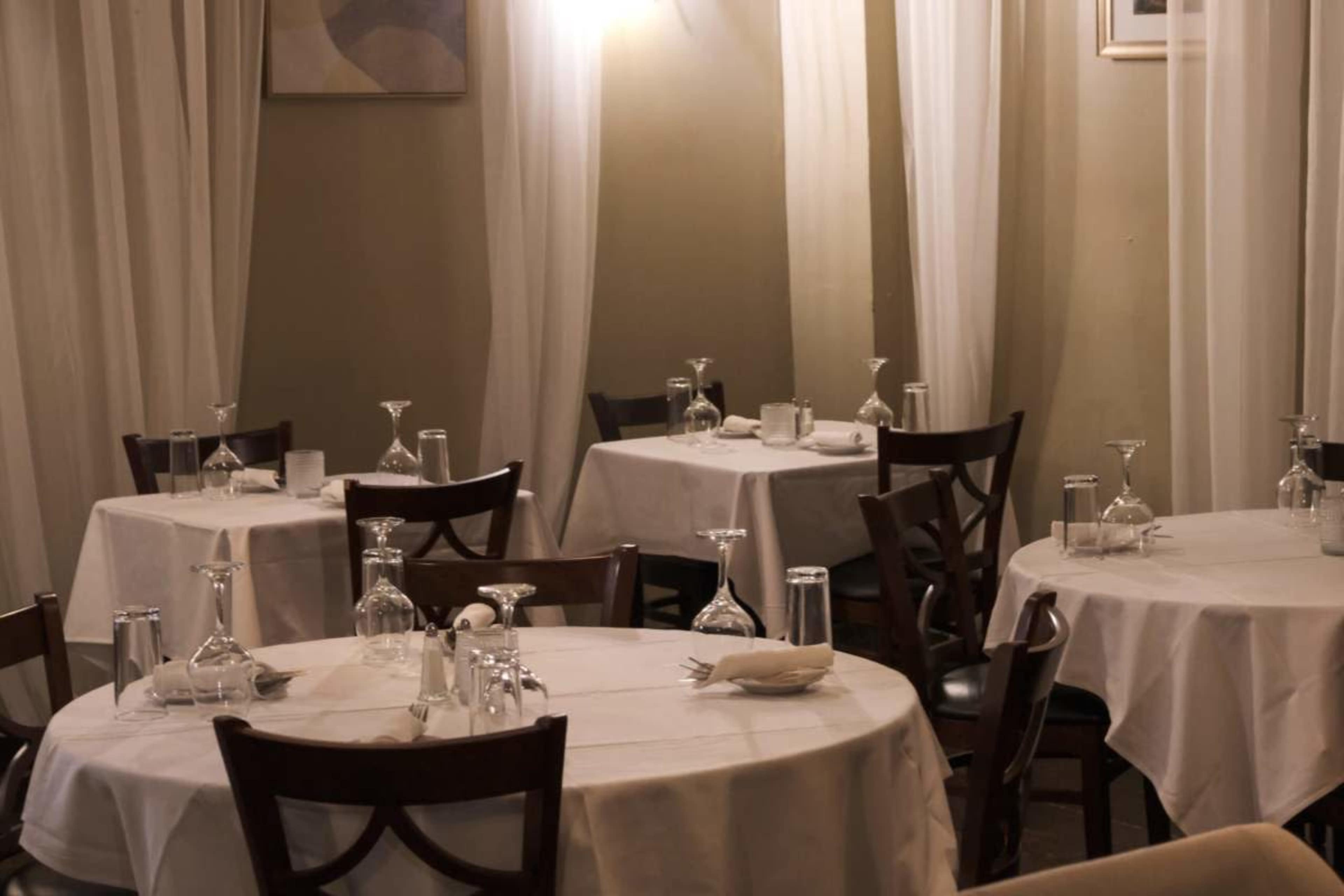 The image shows an empty restaurant interior with round tables covered in white tablecloths, glassware, and chairs arranged around them, surrounded by sheer curtains.