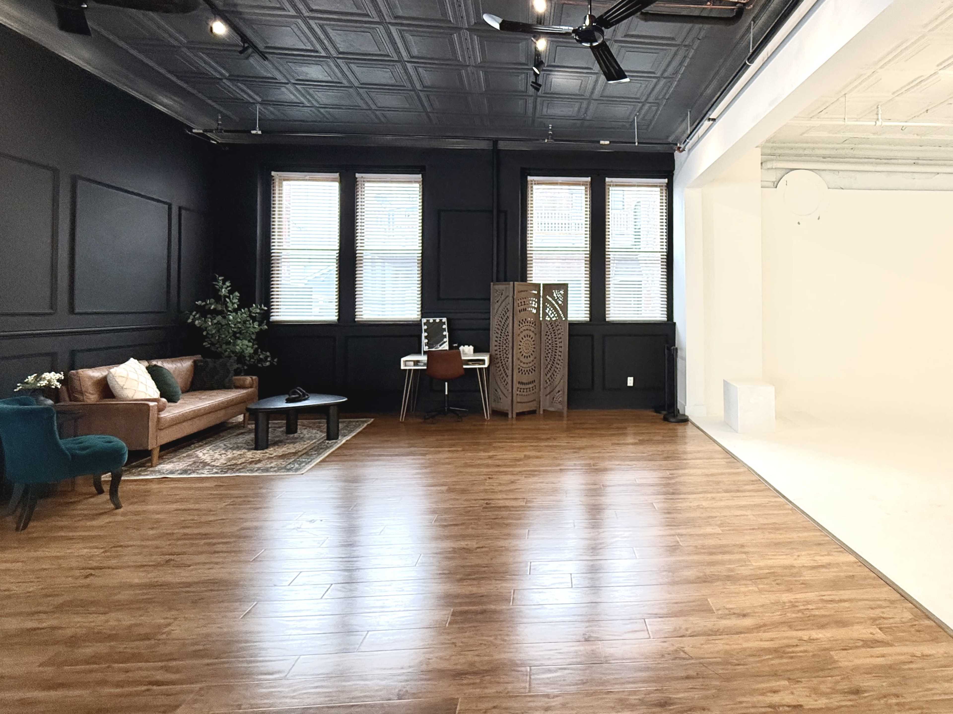 The image shows a spacious interior with wooden flooring featuring a brown sofa, a teal chair, a black coffee table, and an office desk beside a decorative room divider, set against a backdrop of windows and contrasting wall colors.