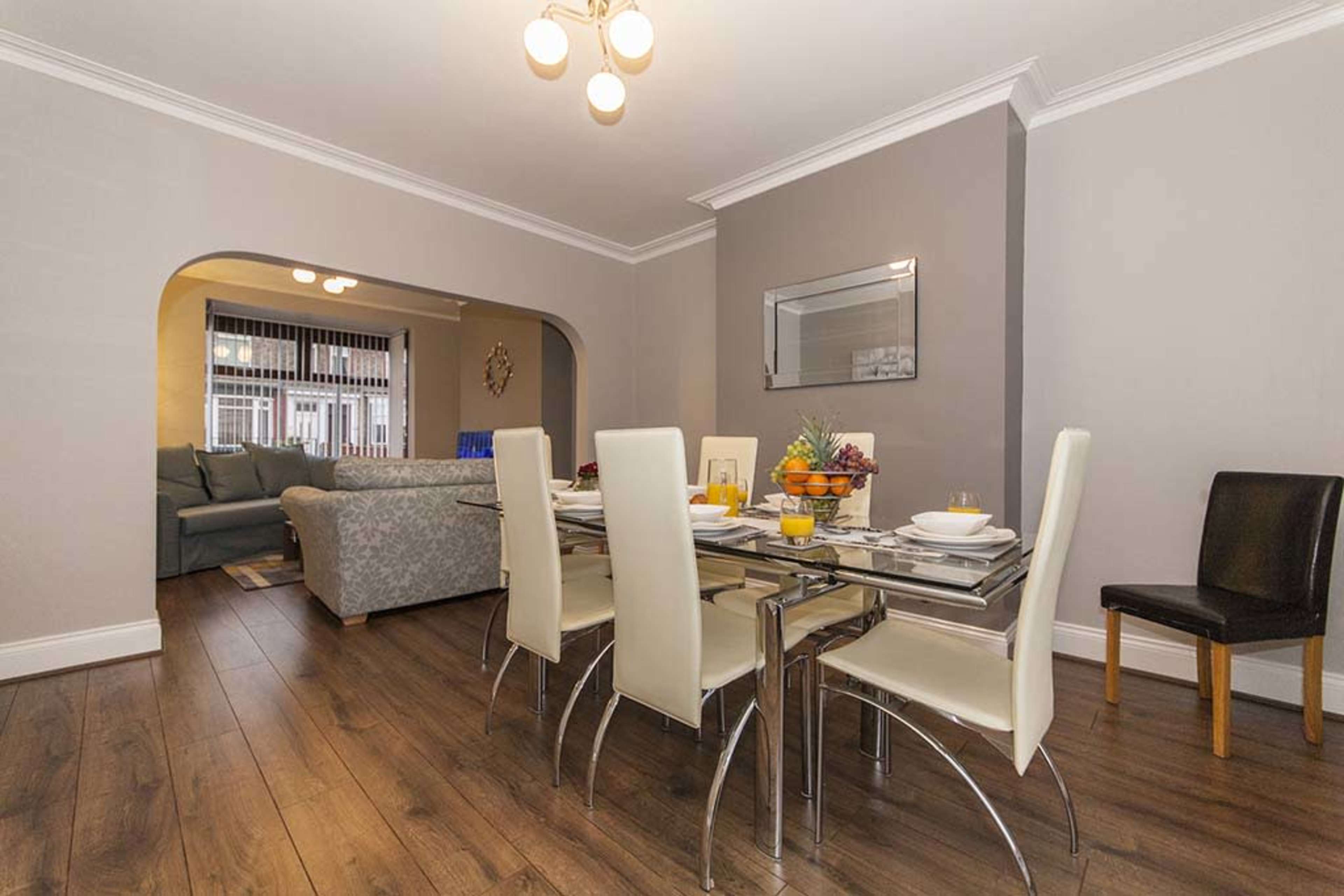 A dining area with a glass table surrounded by white chairs, leading into a living room with a gray sofa in the background.