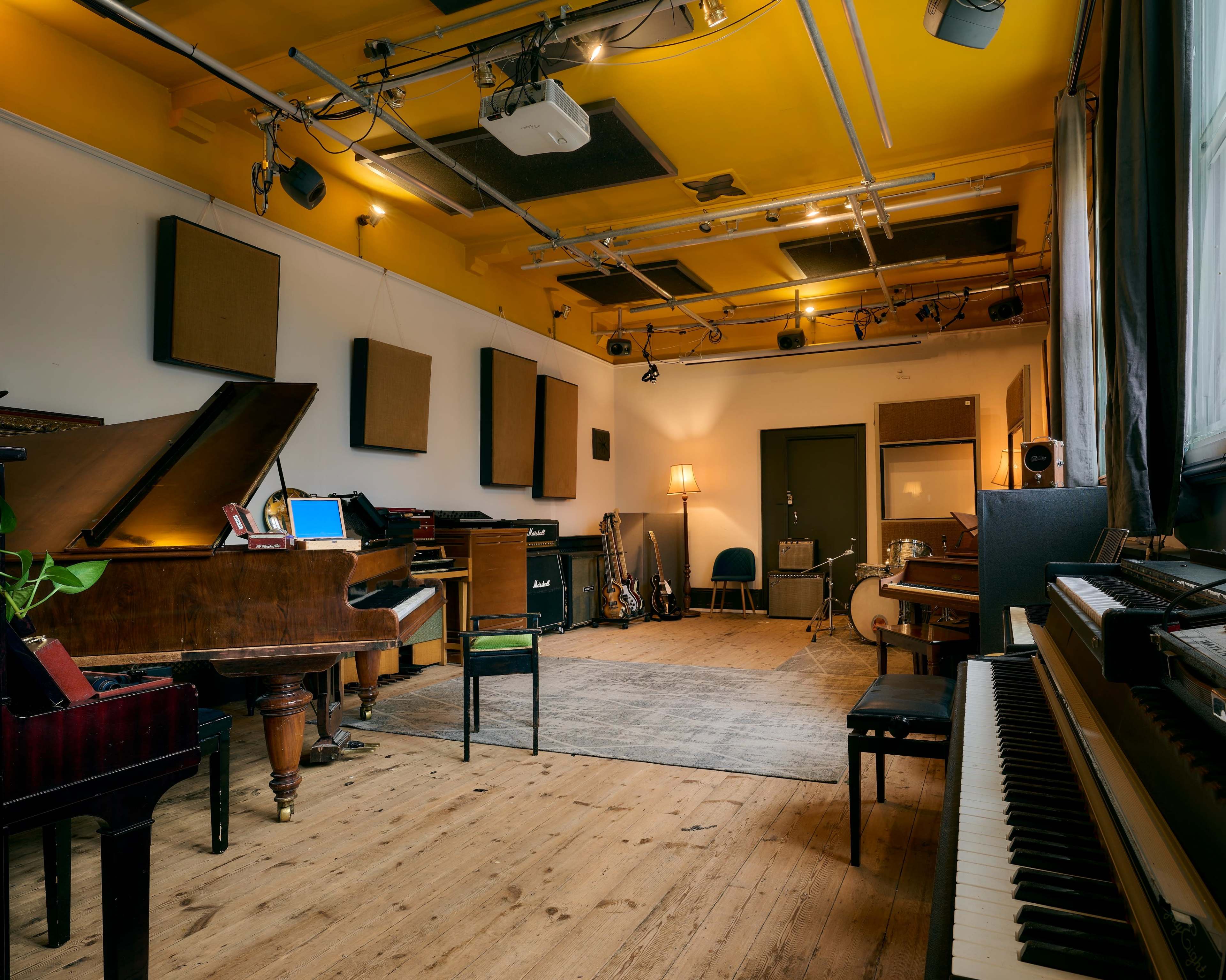 The image shows a music studio filled with various instruments, including two pianos, a guitar, and soundproofing panels on the walls and ceiling.