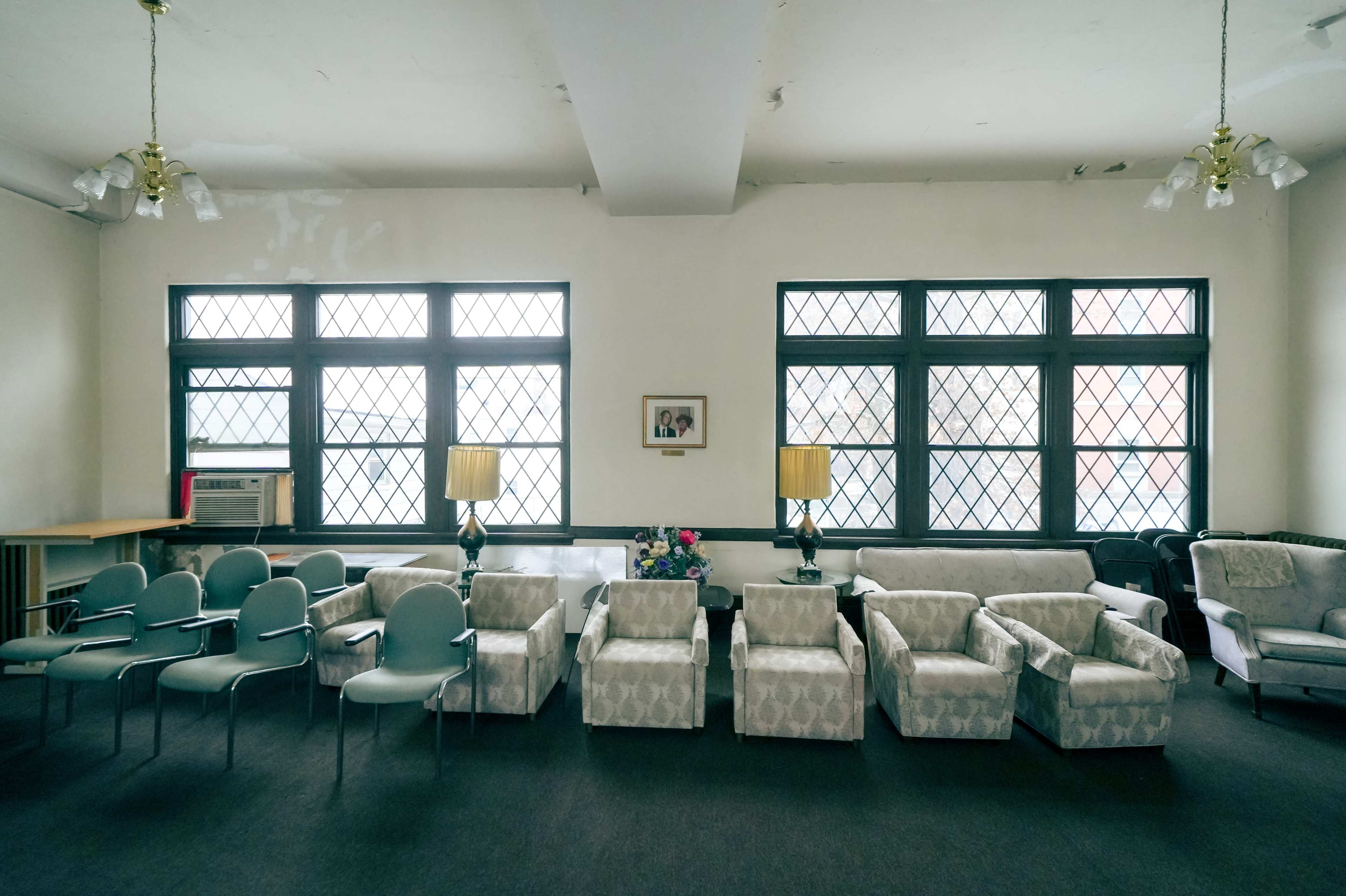 A spacious room with several chairs and armchairs arranged in two rows facing each other, complemented by two lamps and a framed picture on the wall.