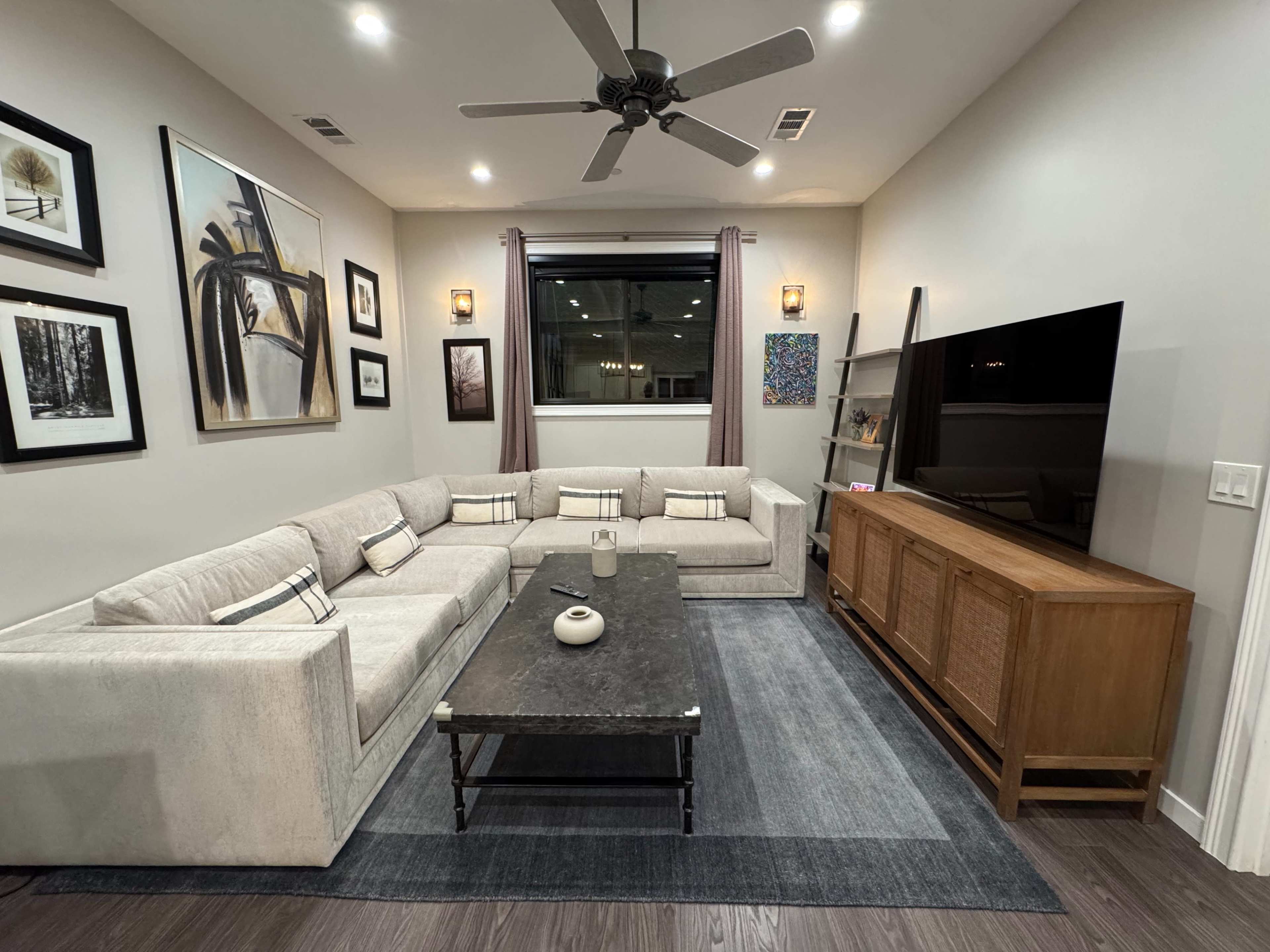The image shows a modern living room with a large sectional sofa, a coffee table, a media console with a television, and framed artworks on the walls.