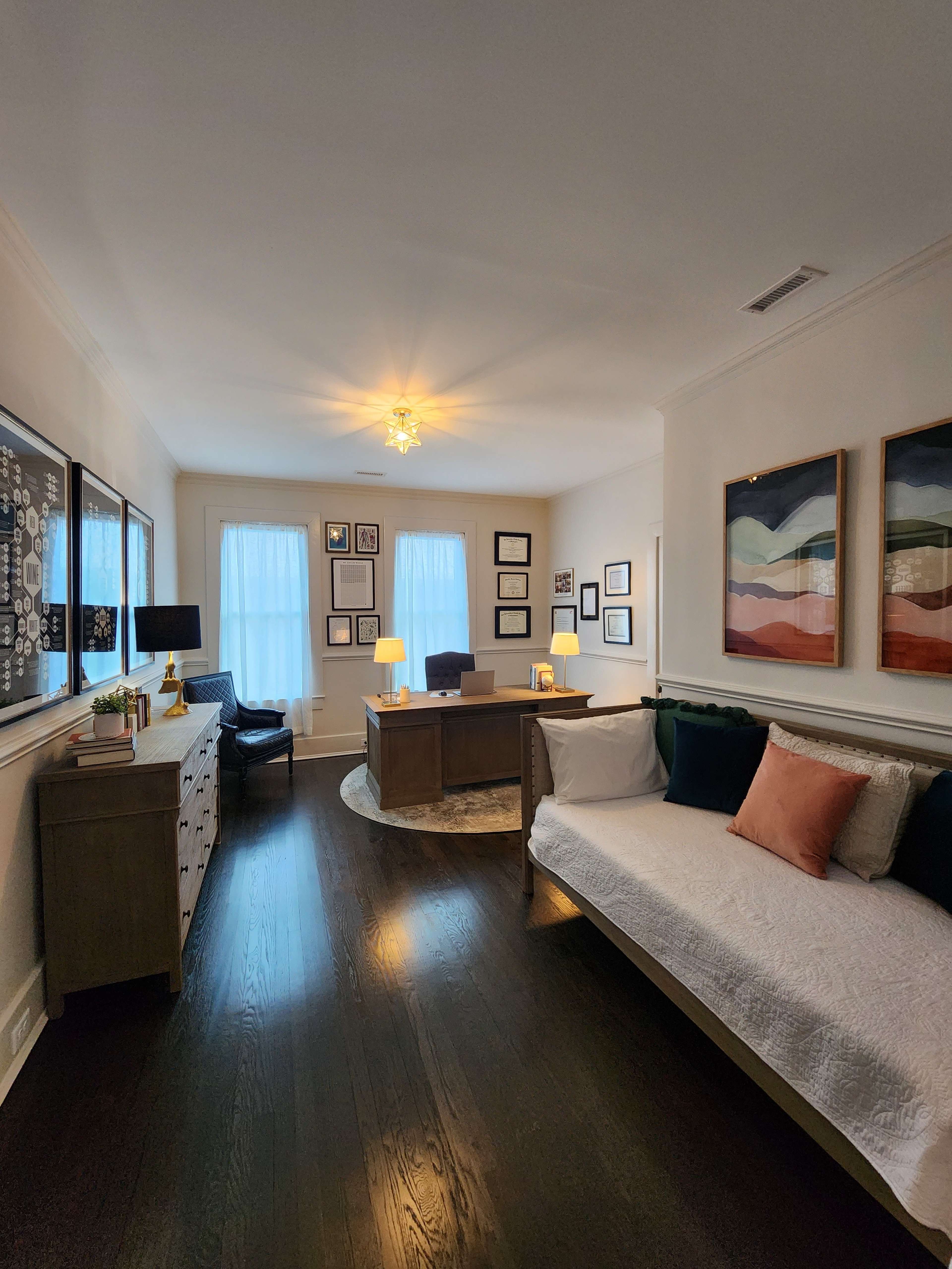 A bright, organized office features a desk at the back, framed artwork on the walls, and a seating area with a white daybed and cushions.
