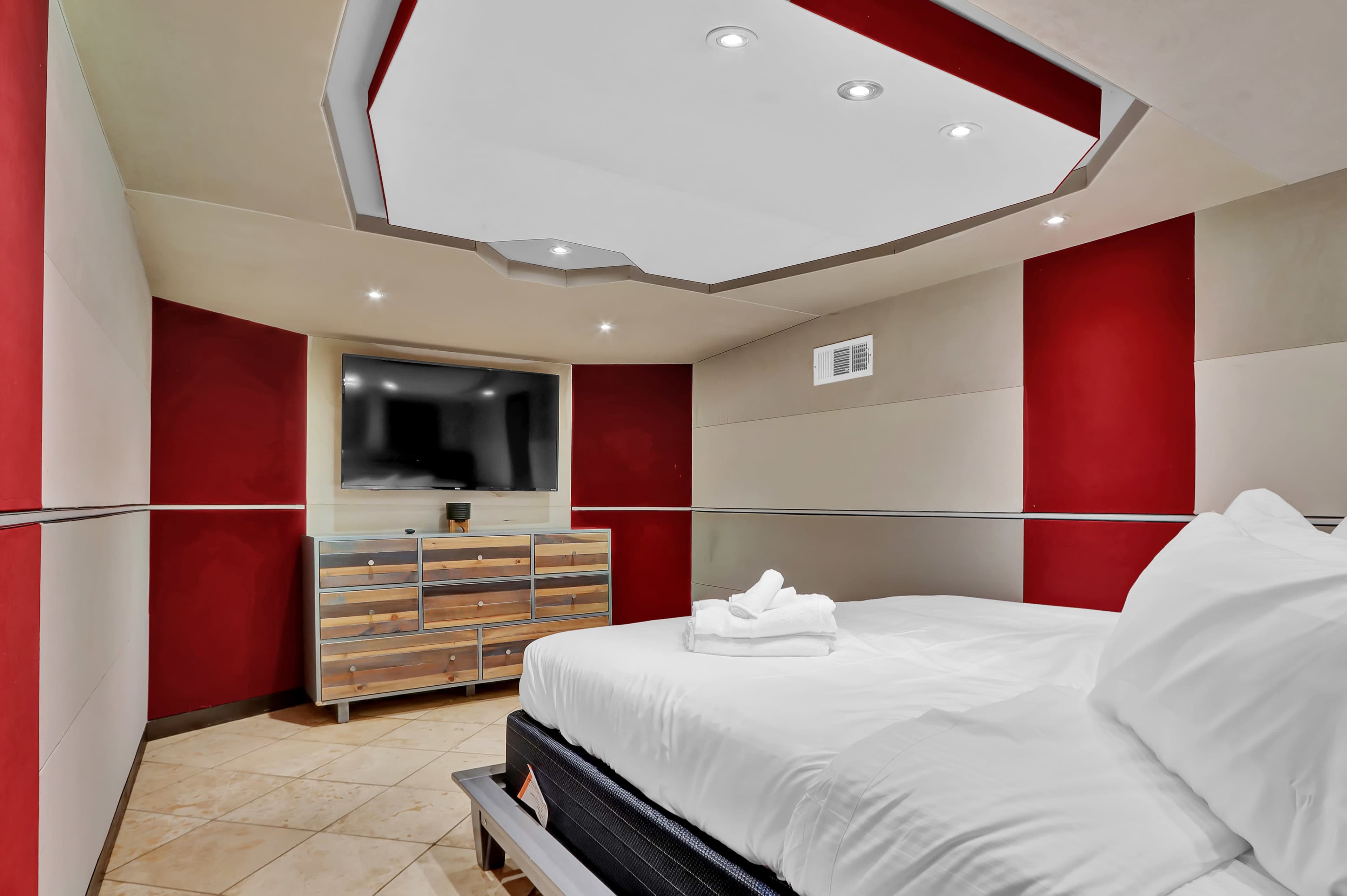 A modern bedroom features a bed with white linens, a flat-screen TV on the wall, and red accent walls.