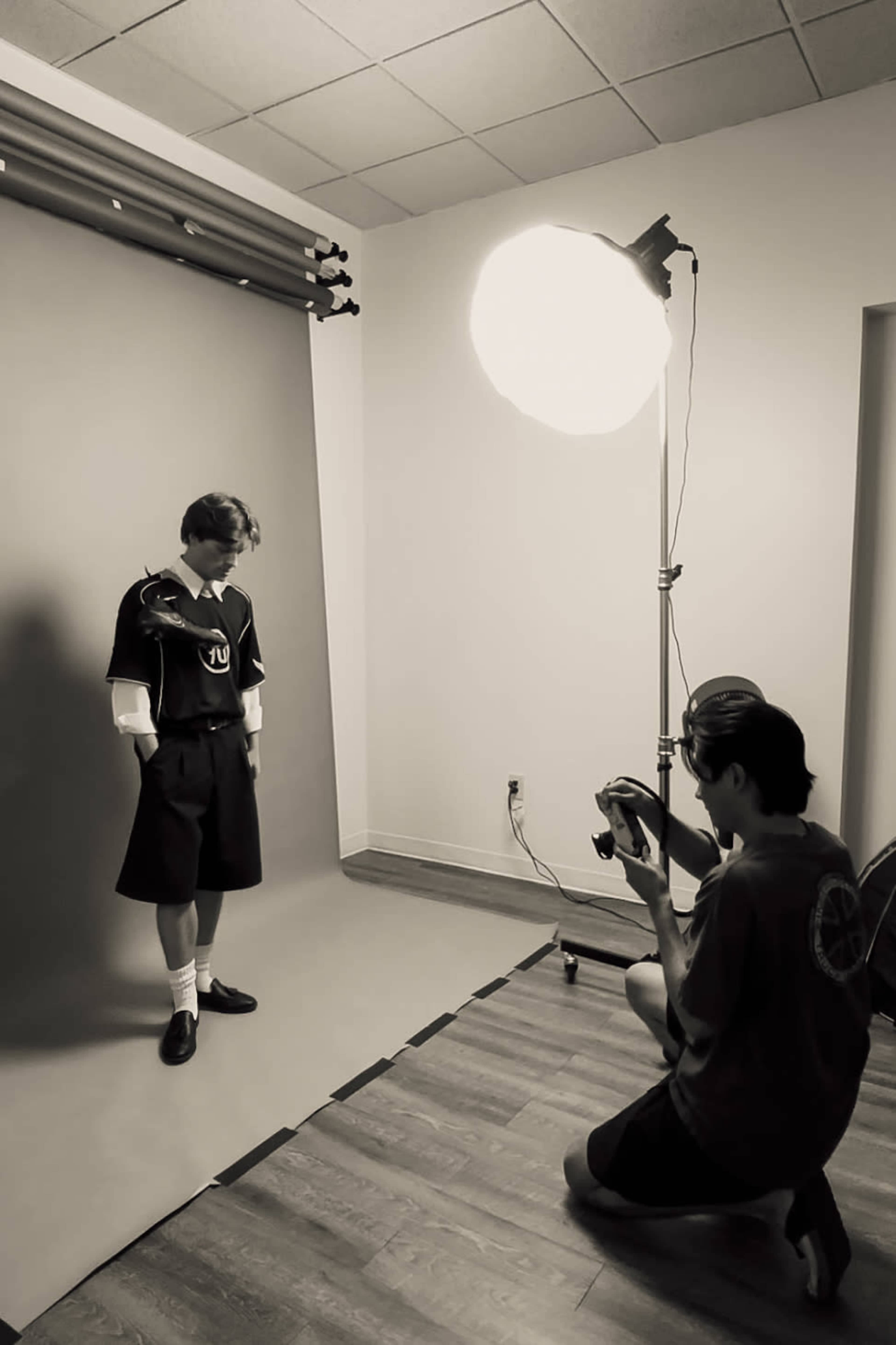 A model stands on a gray backdrop while a photographer kneels and adjusts a camera in a brightly lit studio.