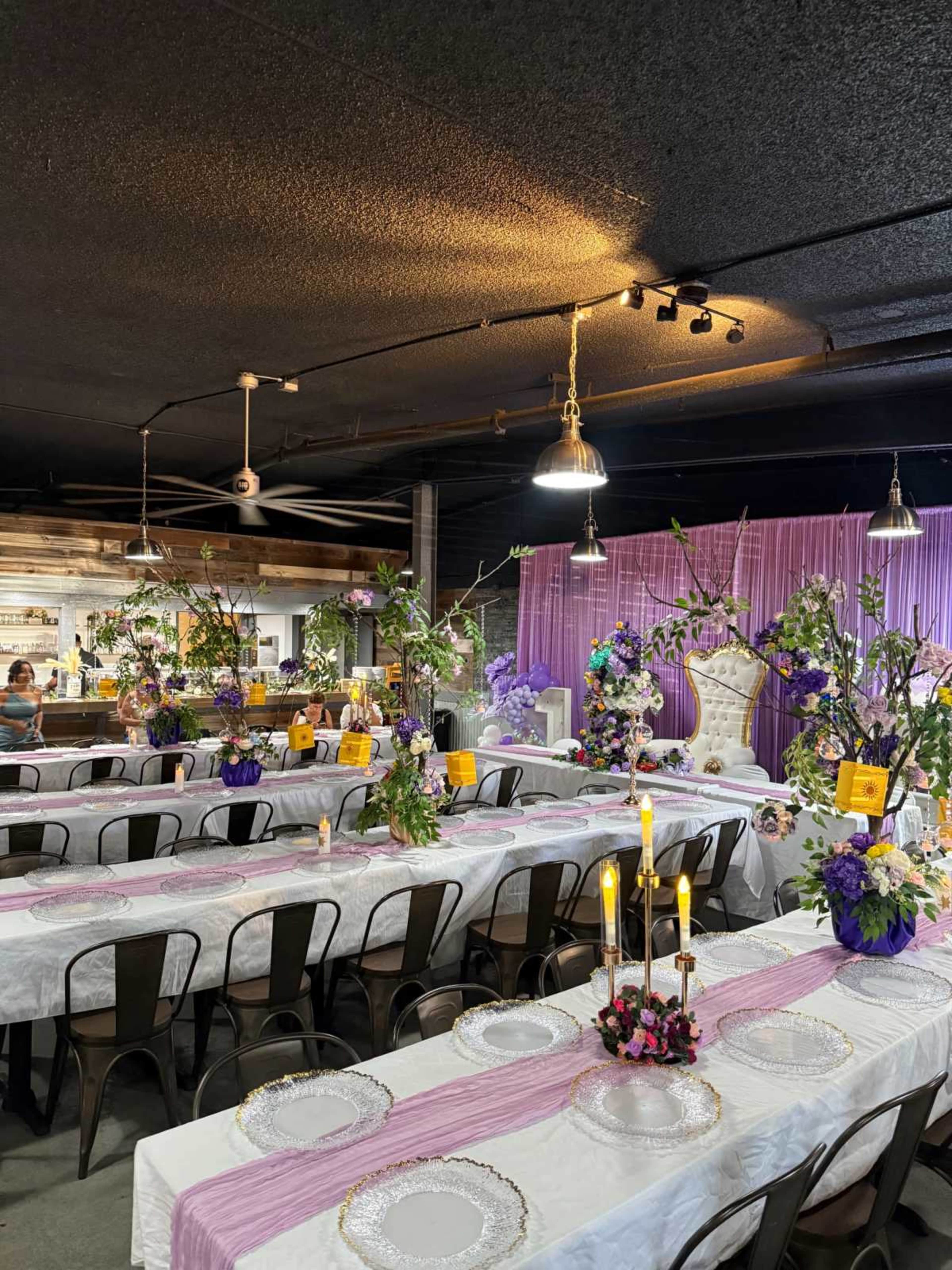The image shows a banquet hall set up with long tables decorated with white tablecloths, floral arrangements, and candles under dim lighting.