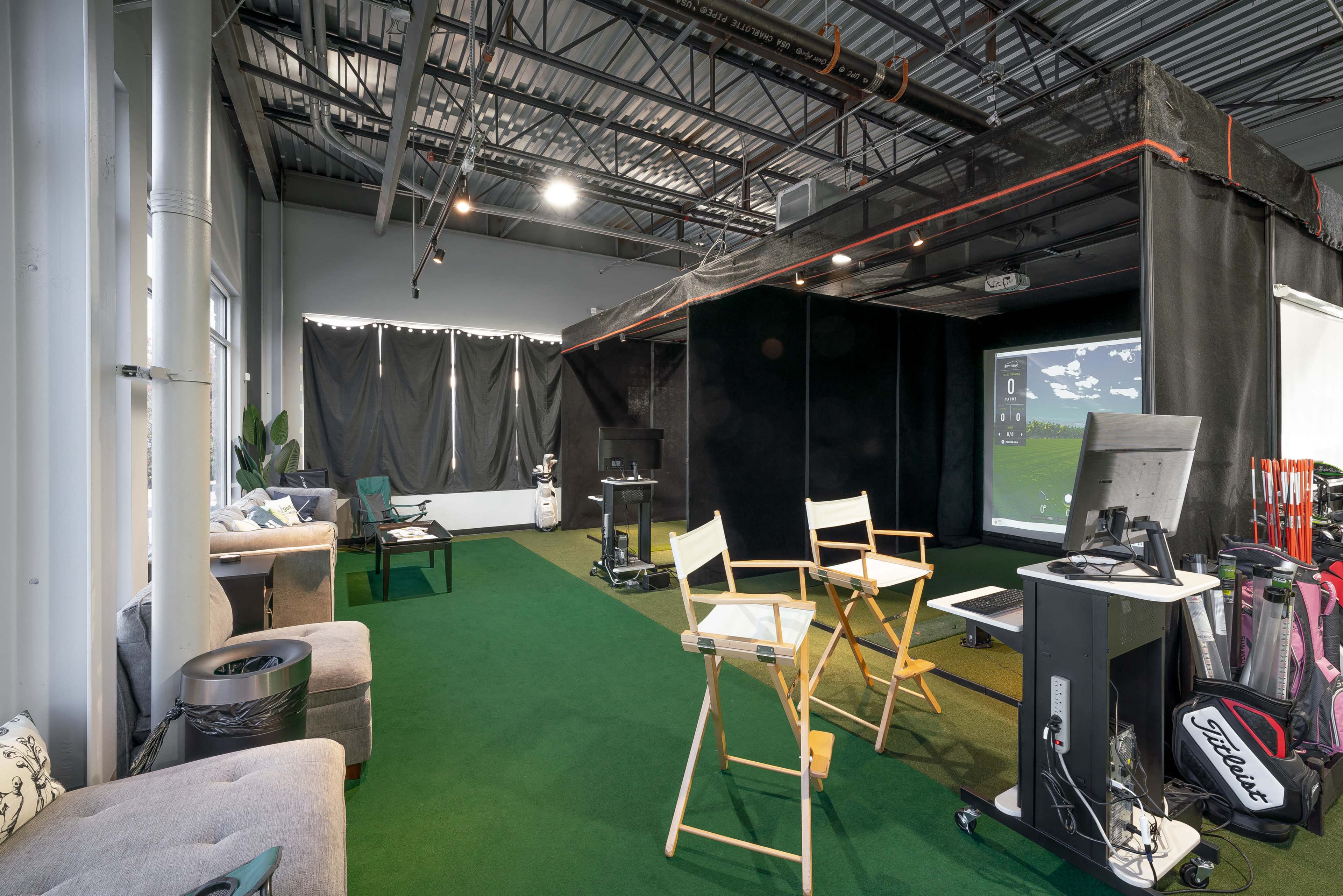 An indoor golf simulation room featuring a green turf floor, seating areas with chairs, a large screen displaying a golf course, and golf bags leaning against a wall.