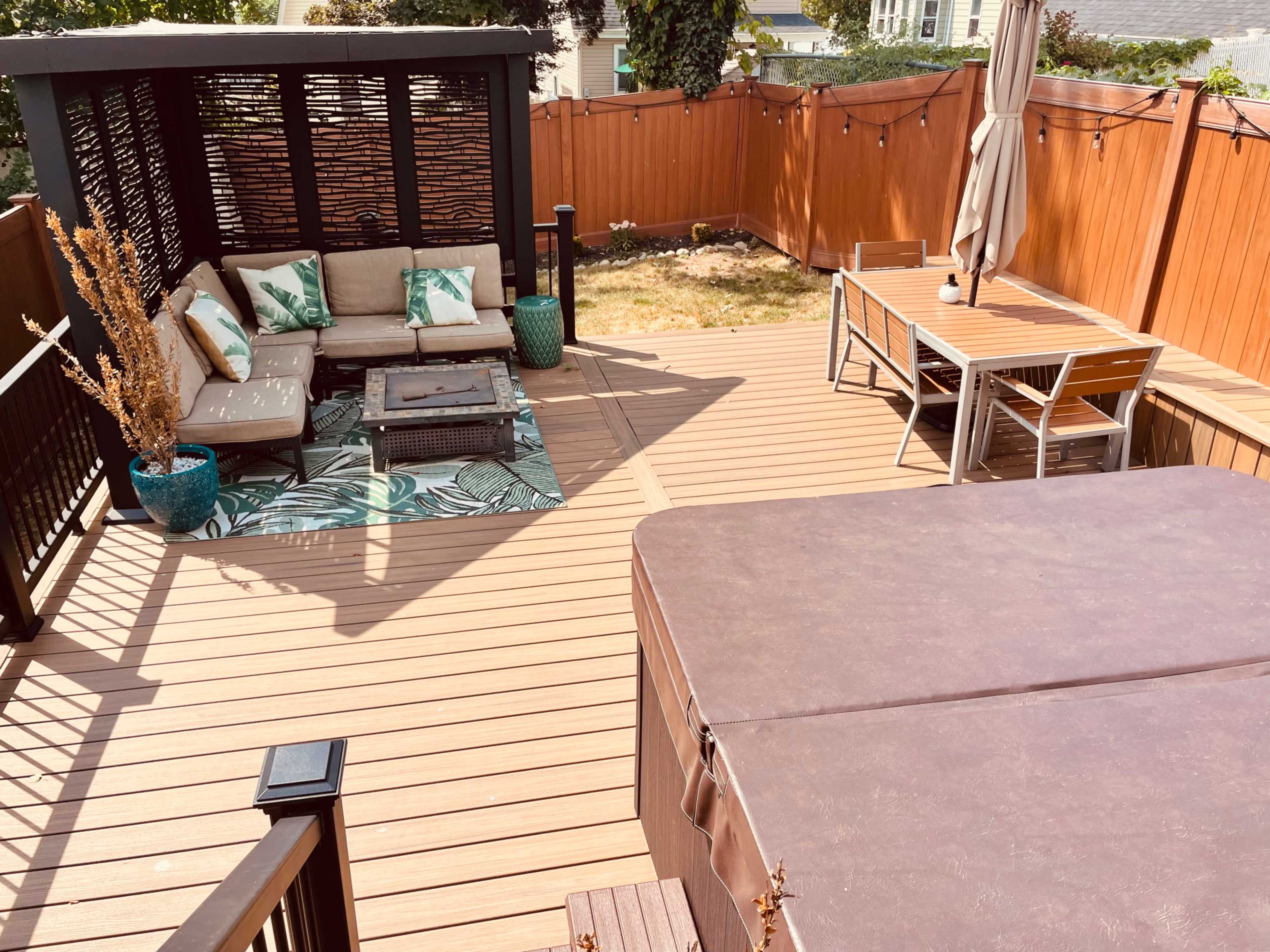The image shows a wooden deck featuring a seating area with a sofa and table, a dining set, a hot tub, and decorative plants, with a fenced backdrop.