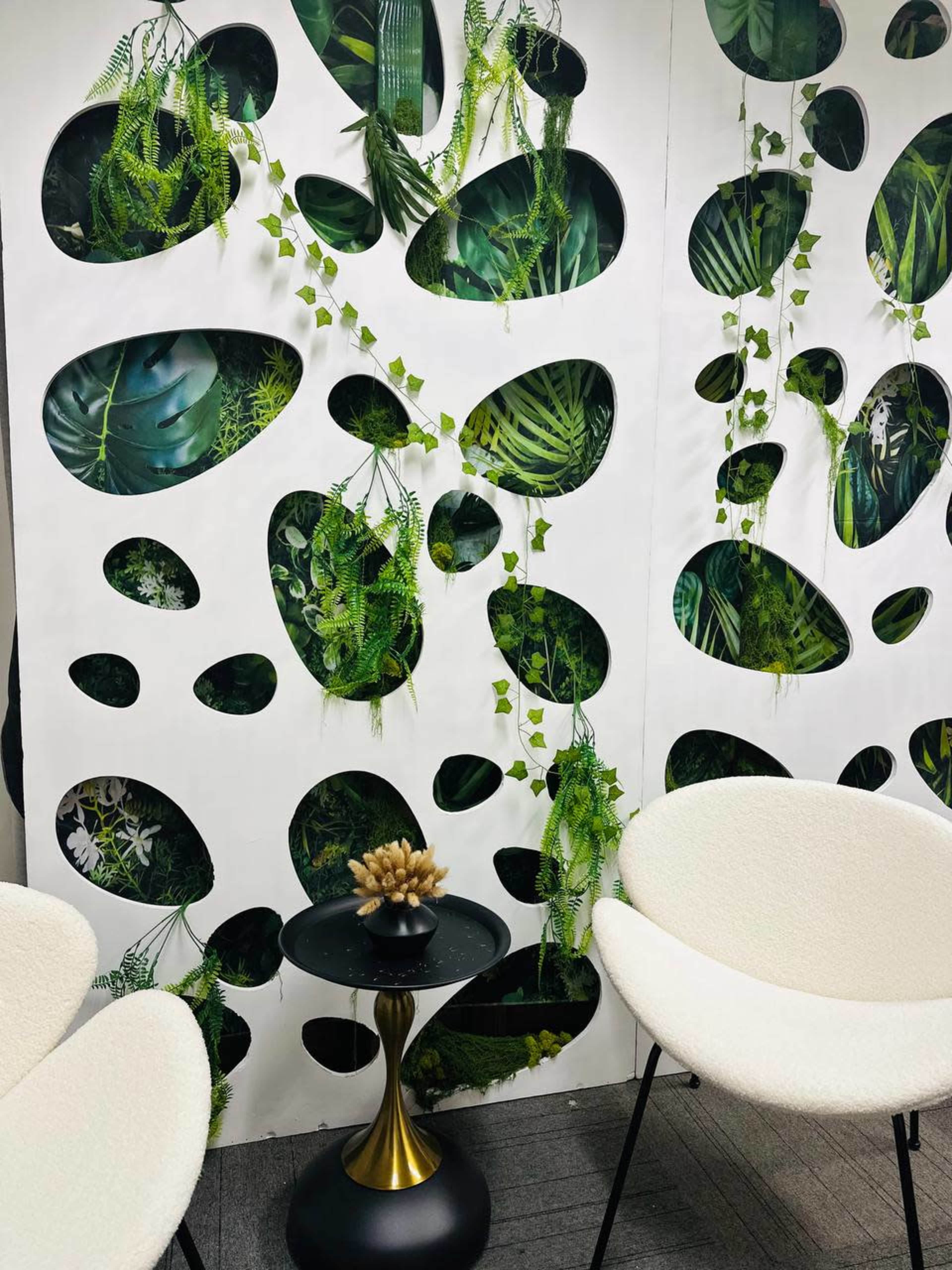 The image shows a modern interior wall featuring cut-out shapes filled with green plants, complemented by two white chairs and a decorative table with a small centerpiece.