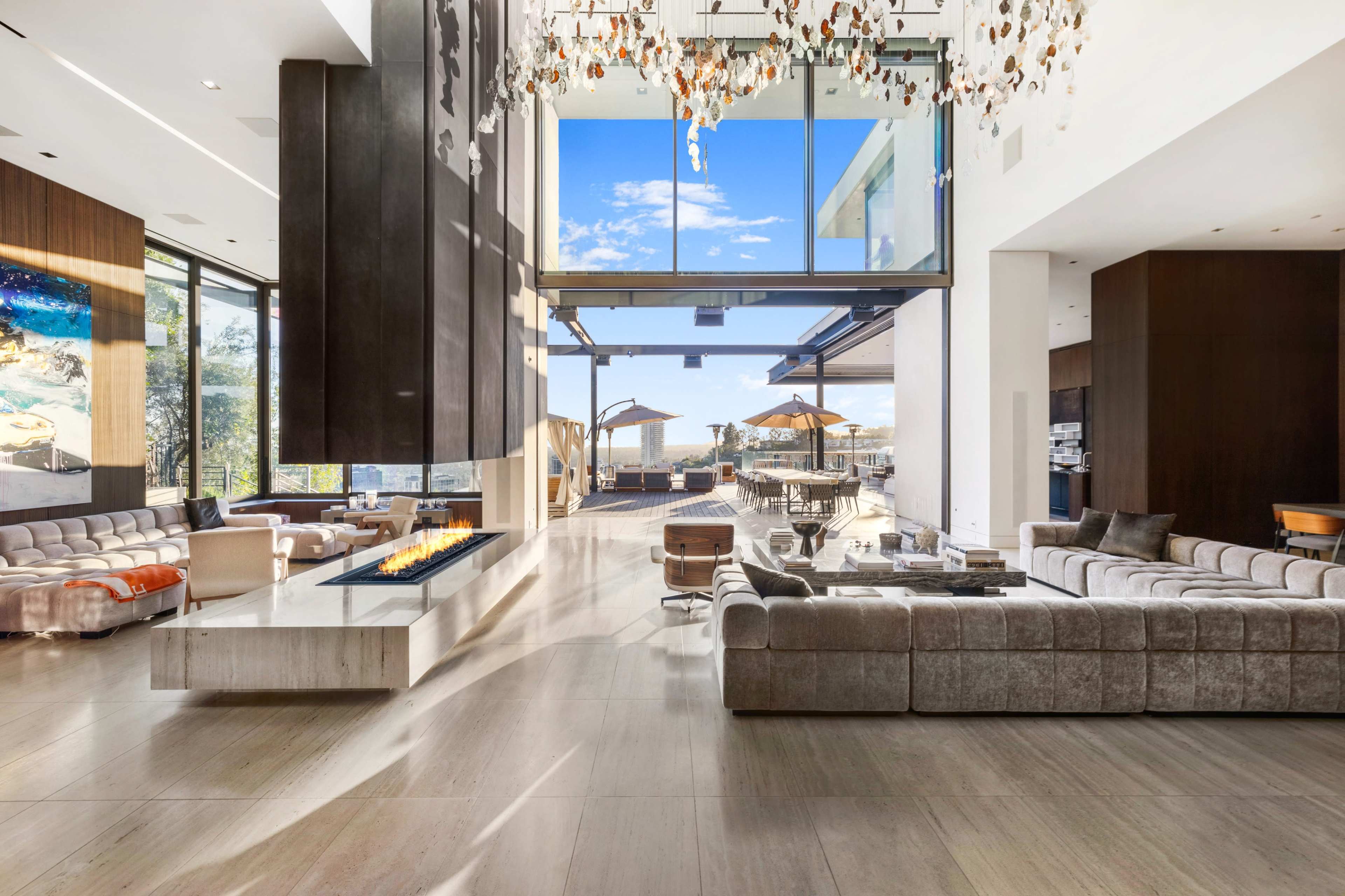 The image shows a spacious modern living area with large windows, a central fireplace, and outdoor seating visible through the glass doors.
