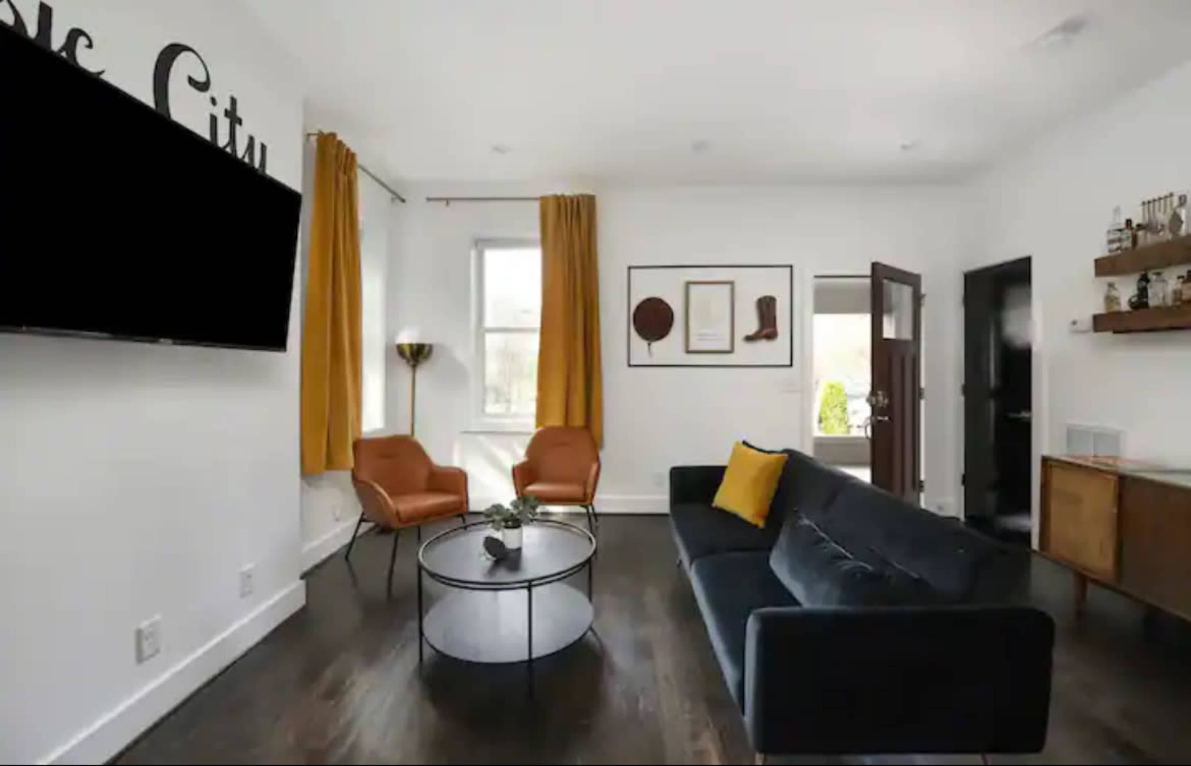 The room features a dark sofa, two orange chairs, a circular coffee table, and a wall-mounted television, with large windows adorned in yellow curtains.