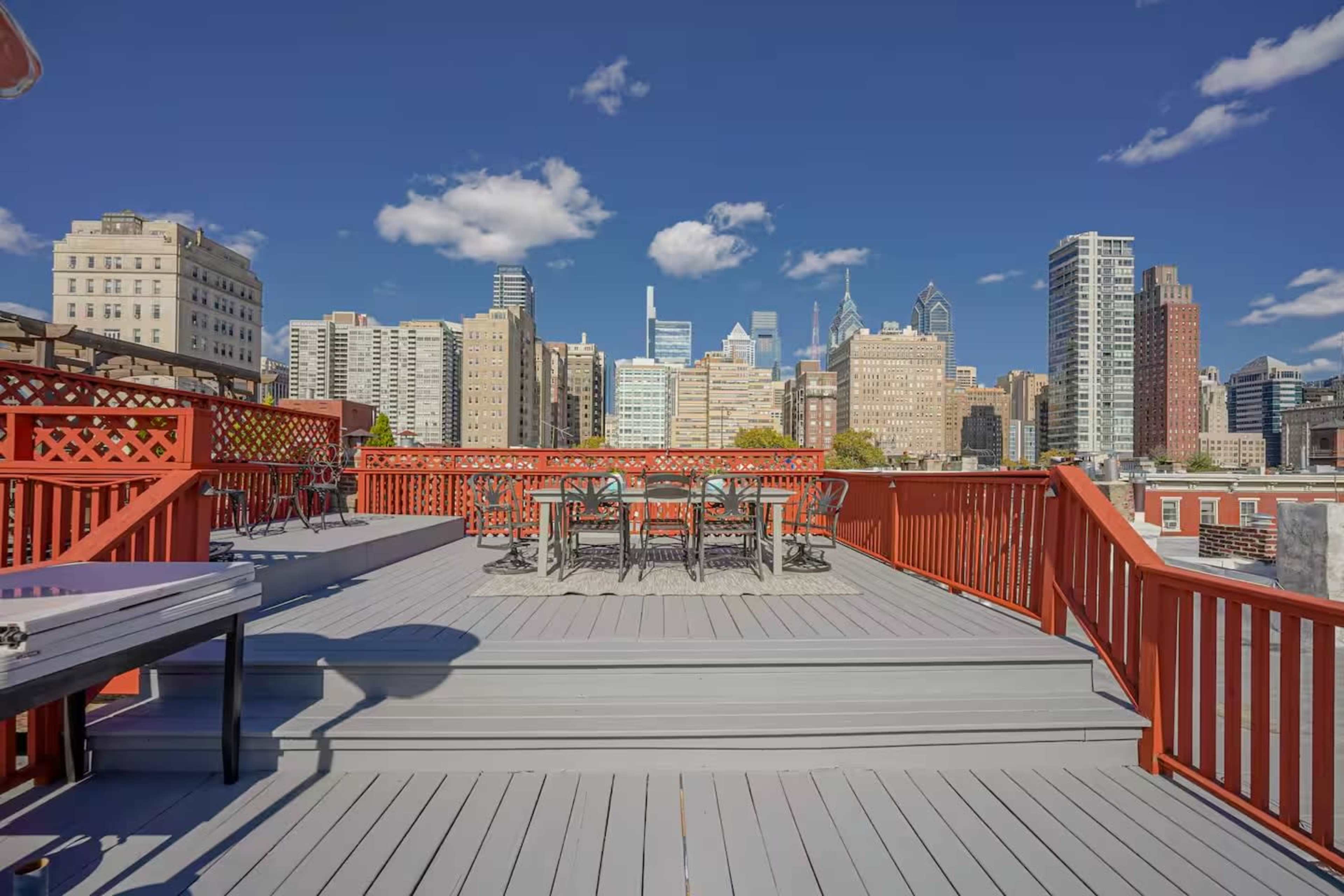 The image shows a rooftop deck with wooden railings and a dining set, overlooking a city skyline on a clear day.