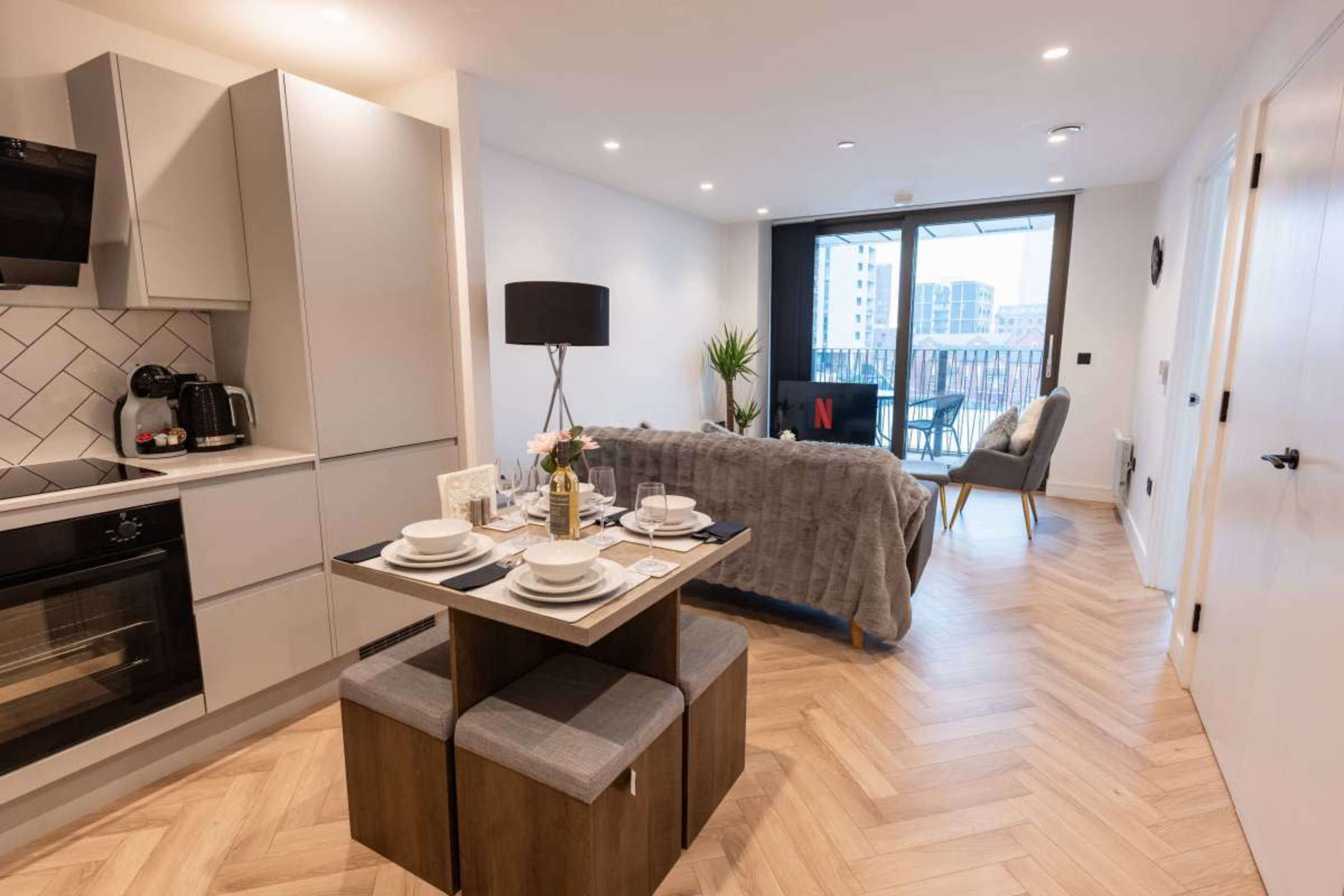 The image shows a compact, modern studio apartment featuring a small dining table with plates, a kitchenette, a bed area, and a balcony visible through large windows.