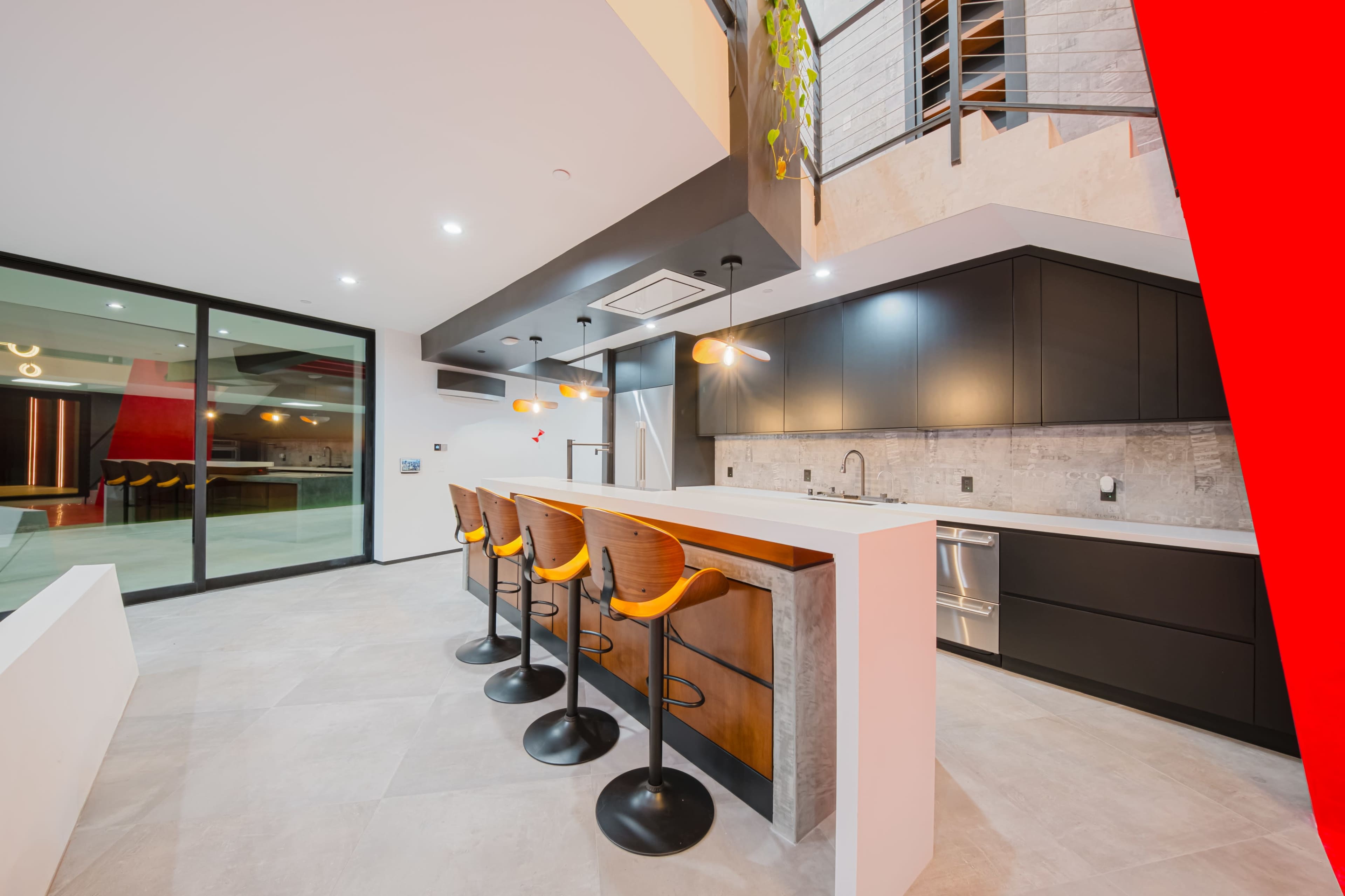 The image shows a modern kitchen with a central island, bar stools, sleek cabinetry, and large windows overlooking an outdoor space.