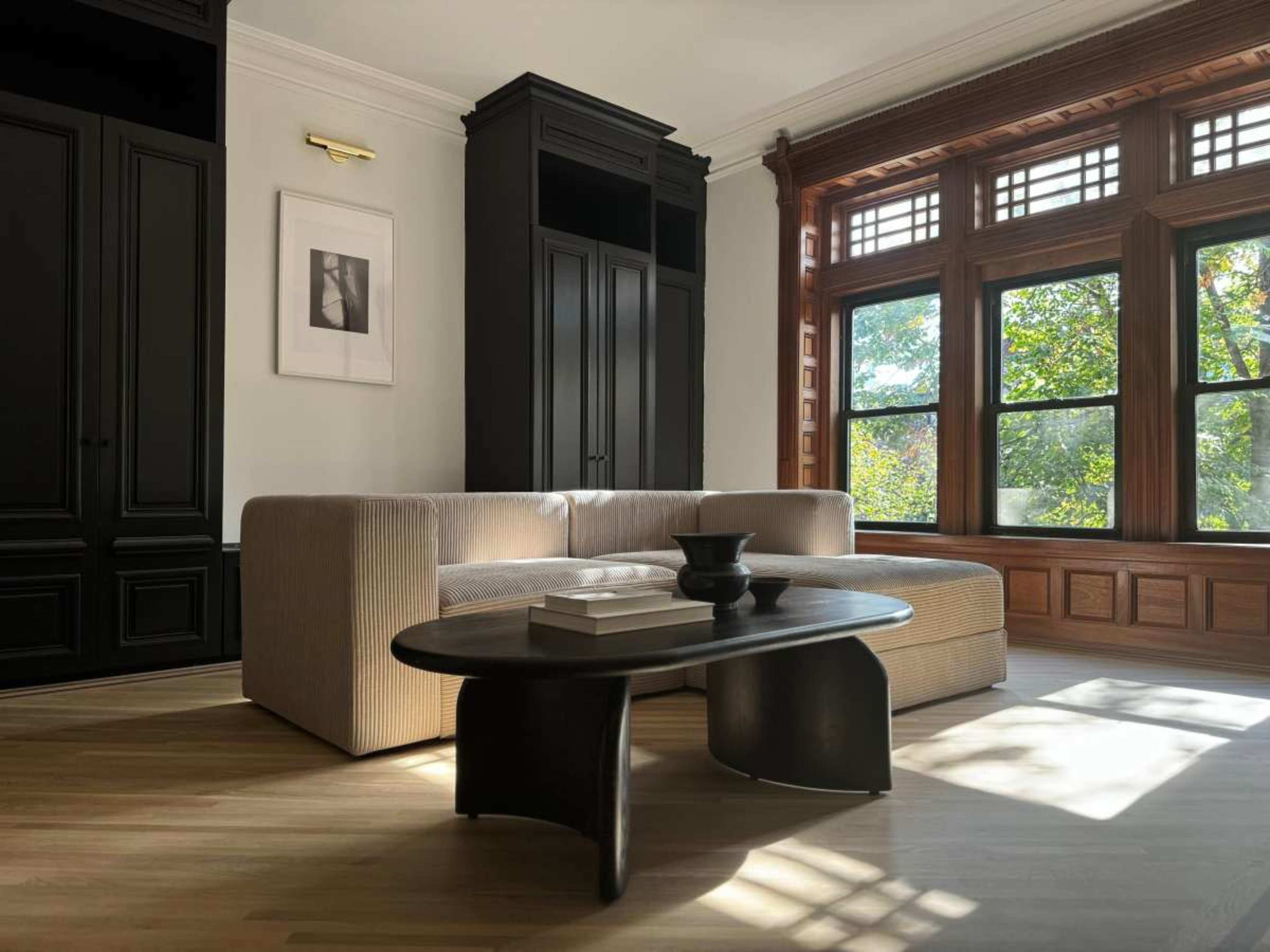 A modern living room features a light-colored sofa, a dark coffee table, and large windows that let in natural light, showcasing wooden trim and cabinetry.