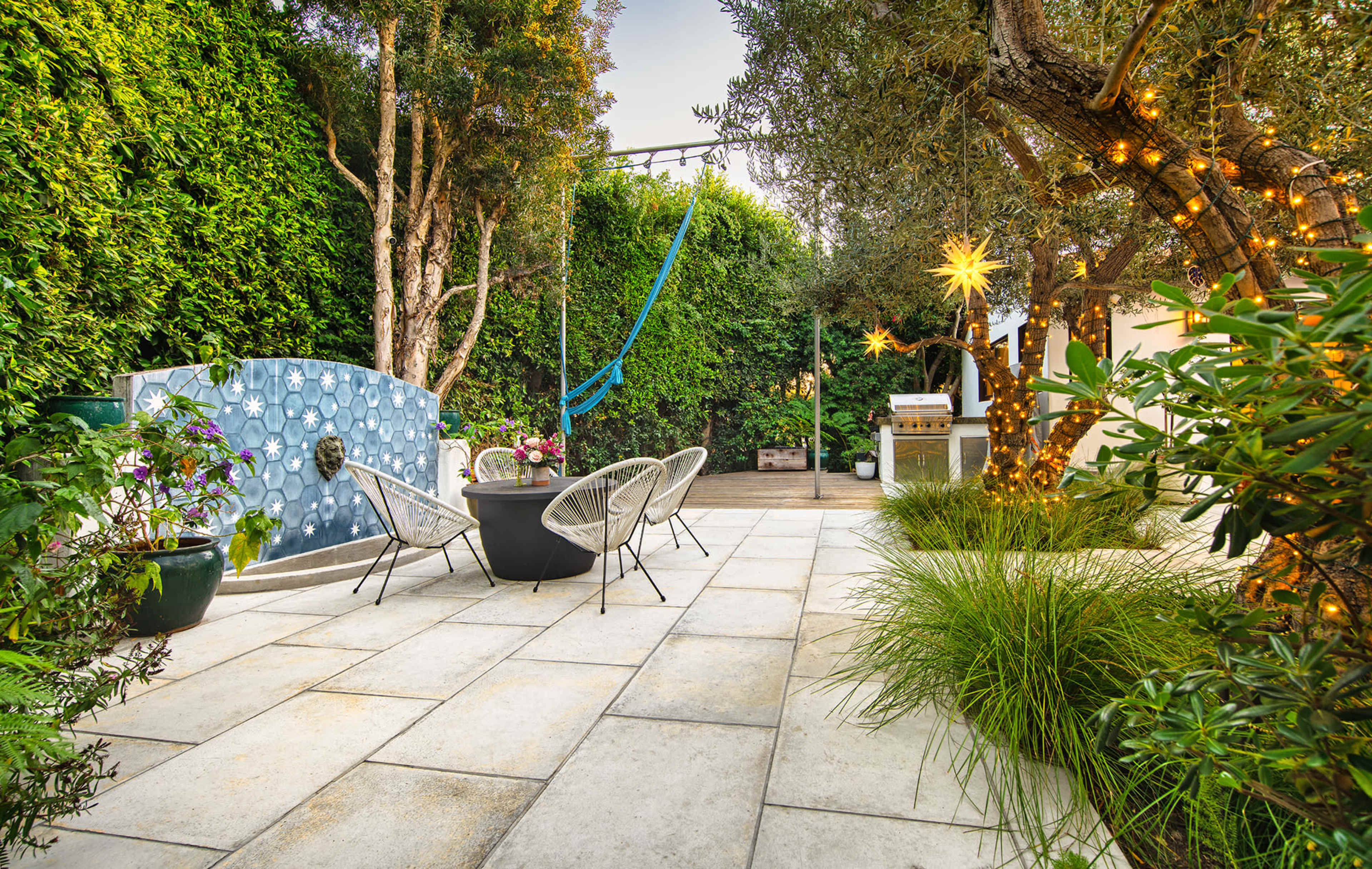 A landscaped outdoor patio area with stone tiles, potted plants, and a blue wall feature, accompanied by a seating arrangement of black chairs and a hammock nearby.