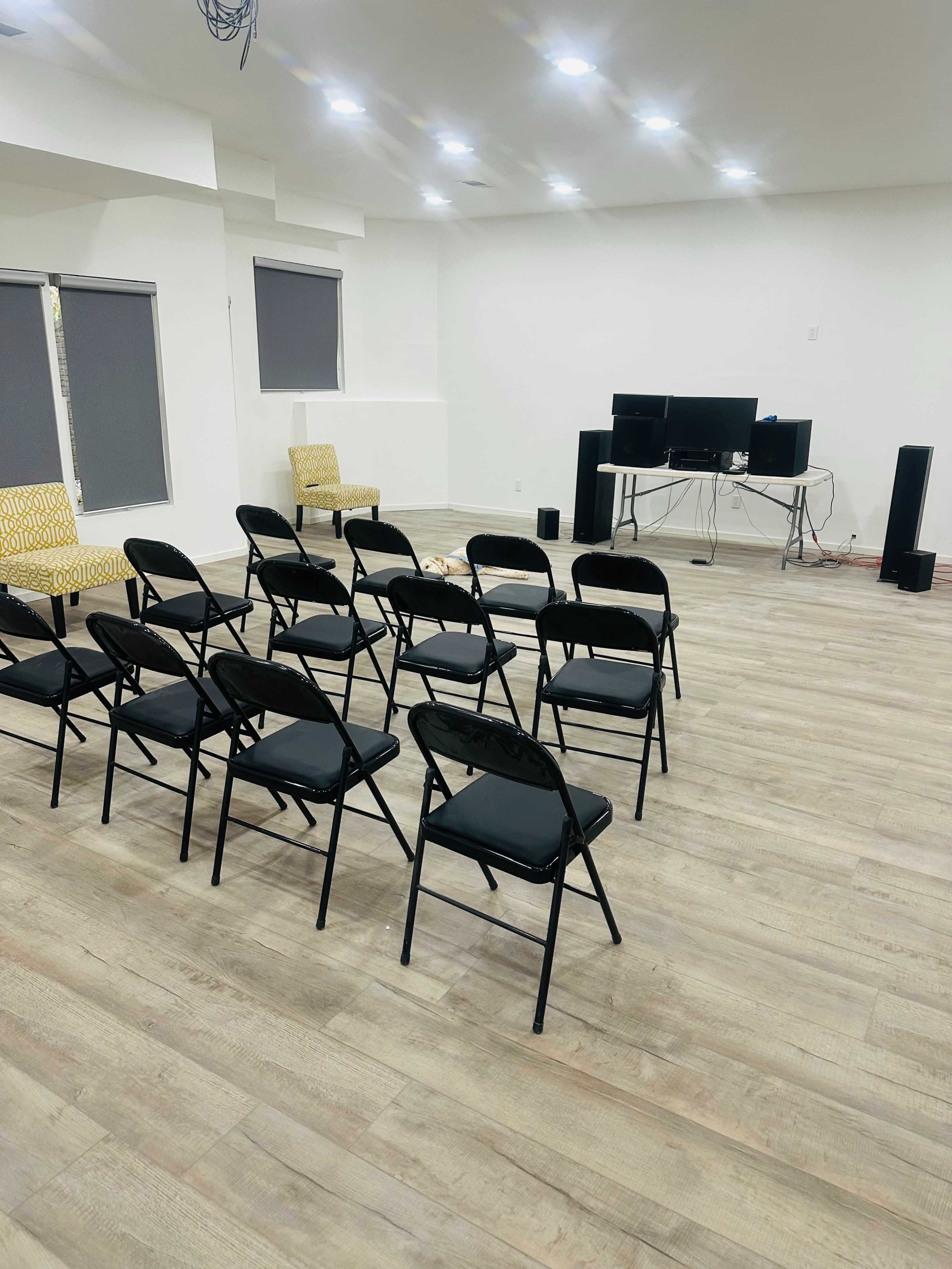 The image shows a spacious room with several black folding chairs arranged in rows facing a table that holds multiple computer monitors and speakers.
