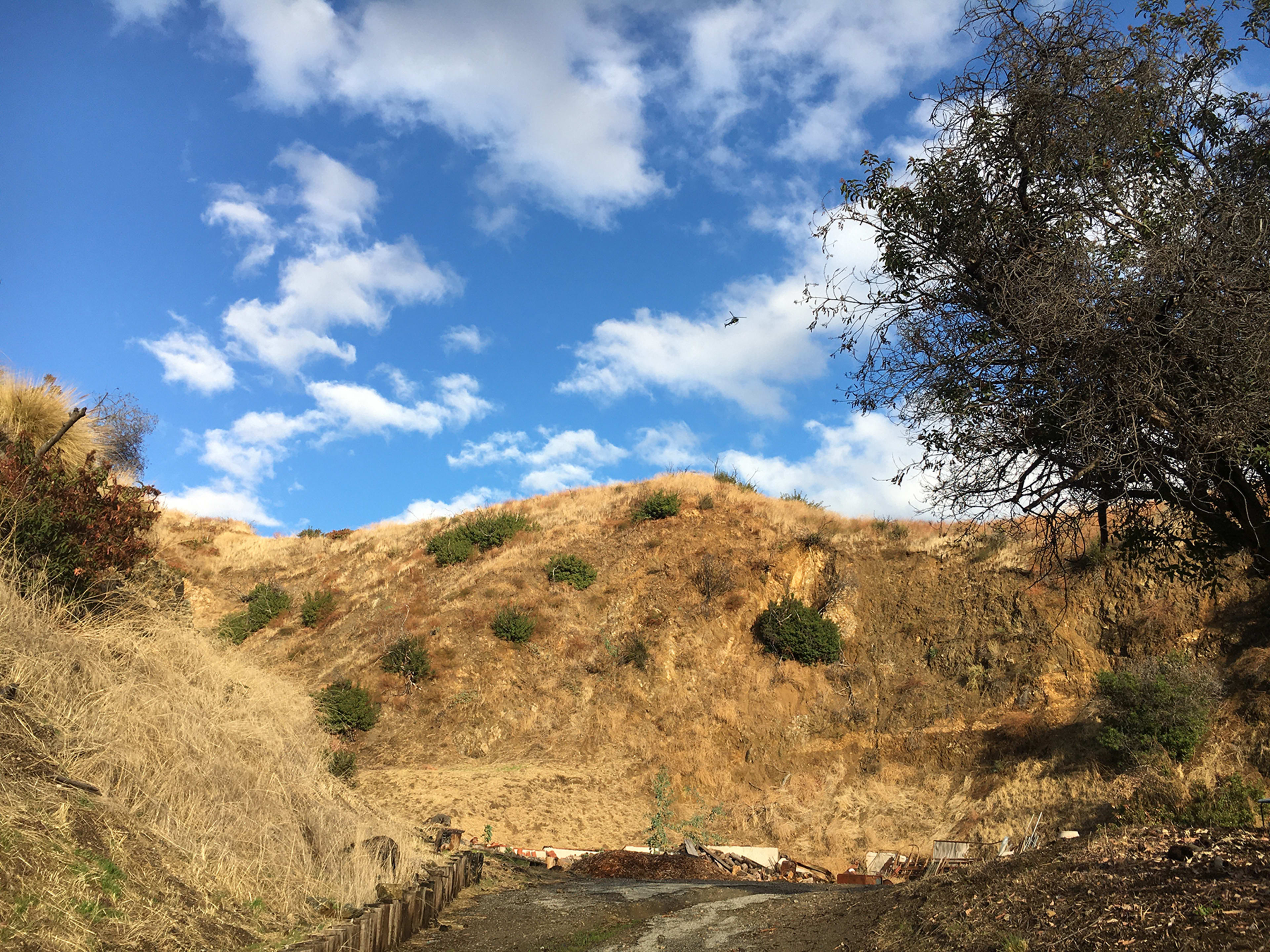 A rugged hillside with dry grass and sparse vegetation is set against a blue sky with scattered clouds.