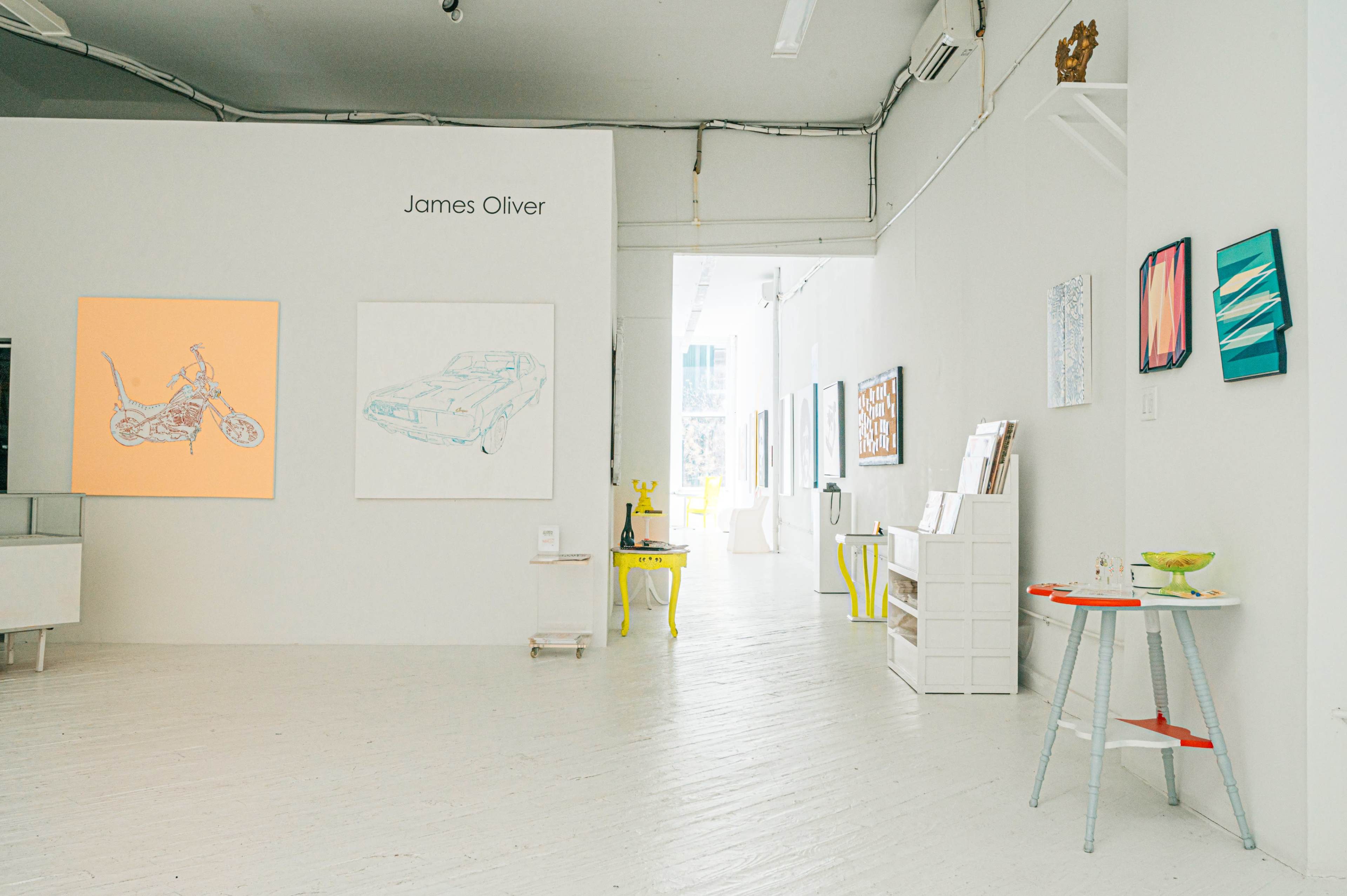 An art gallery with white walls displaying various paintings, including a motorcycle and a car, alongside a small table and chair arrangement.