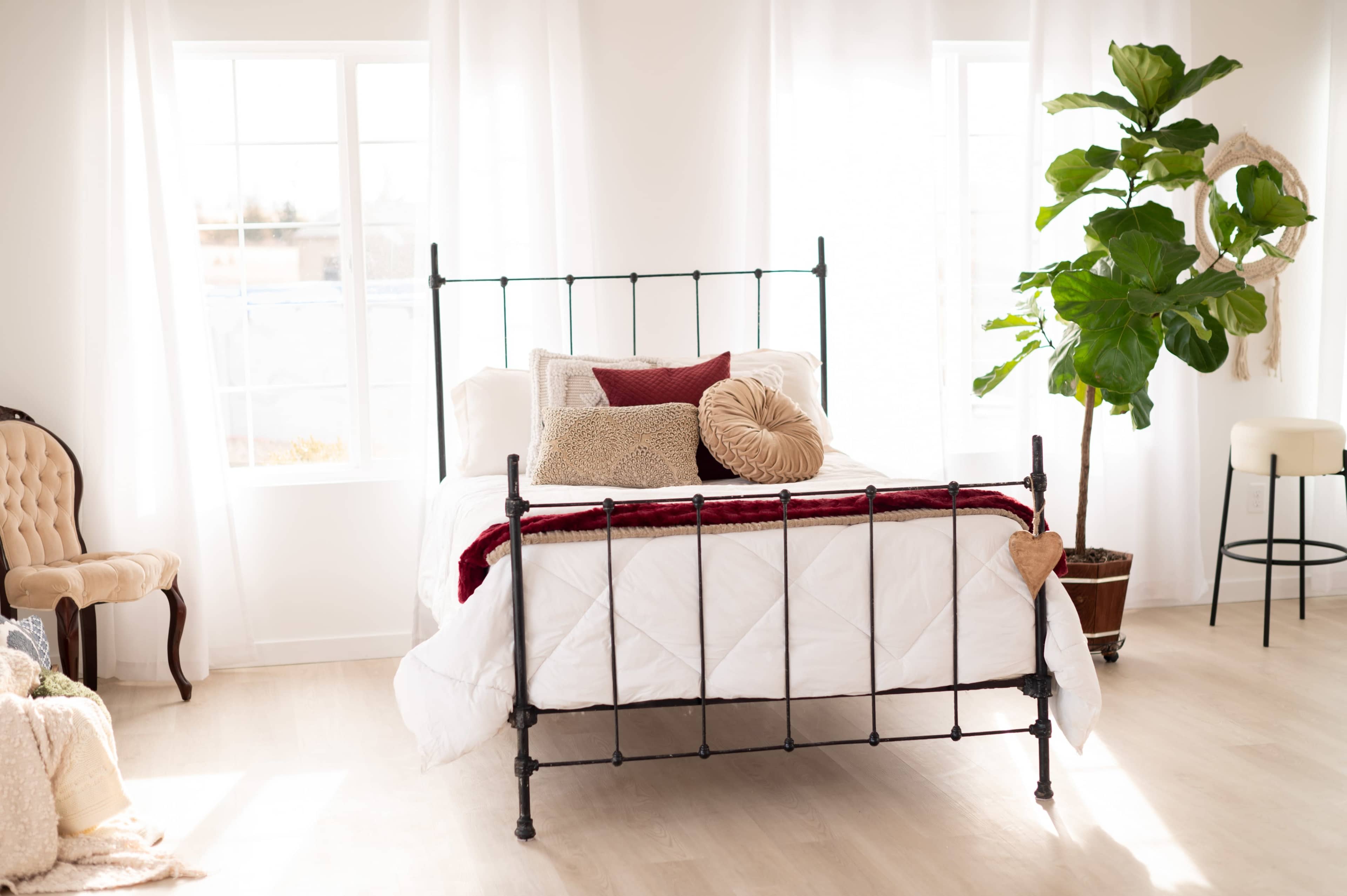 A metal framed bed is centered in a bright, airy room with large windows, accompanied by a chair and a potted plant.