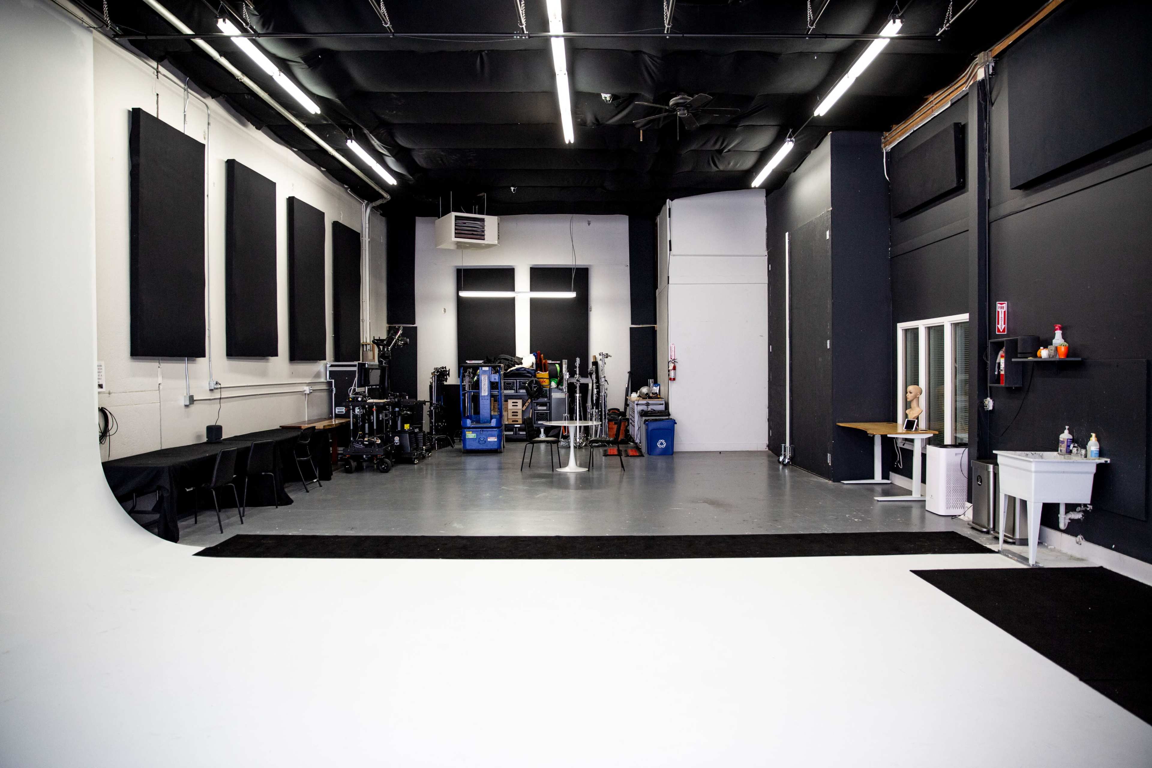 The image shows a spacious photography studio with a white backdrop, black flooring, and various equipment along the walls.