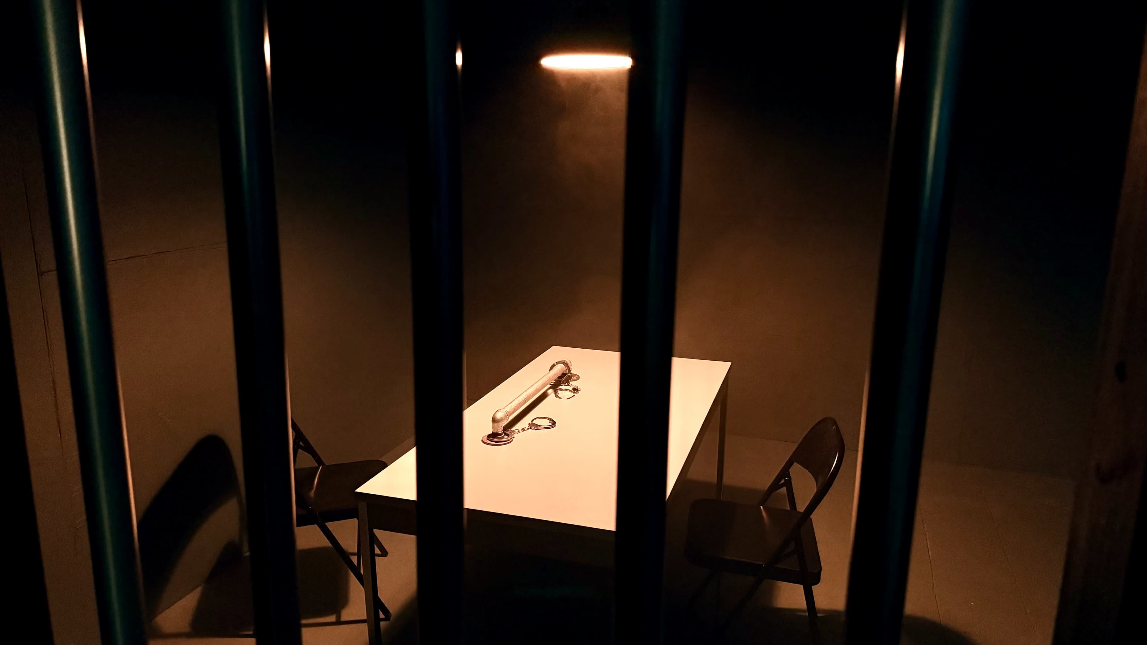 A narrow view shows a dimly lit interrogation room with a table, two chairs, and handcuffs on the table, framed by prison bars.