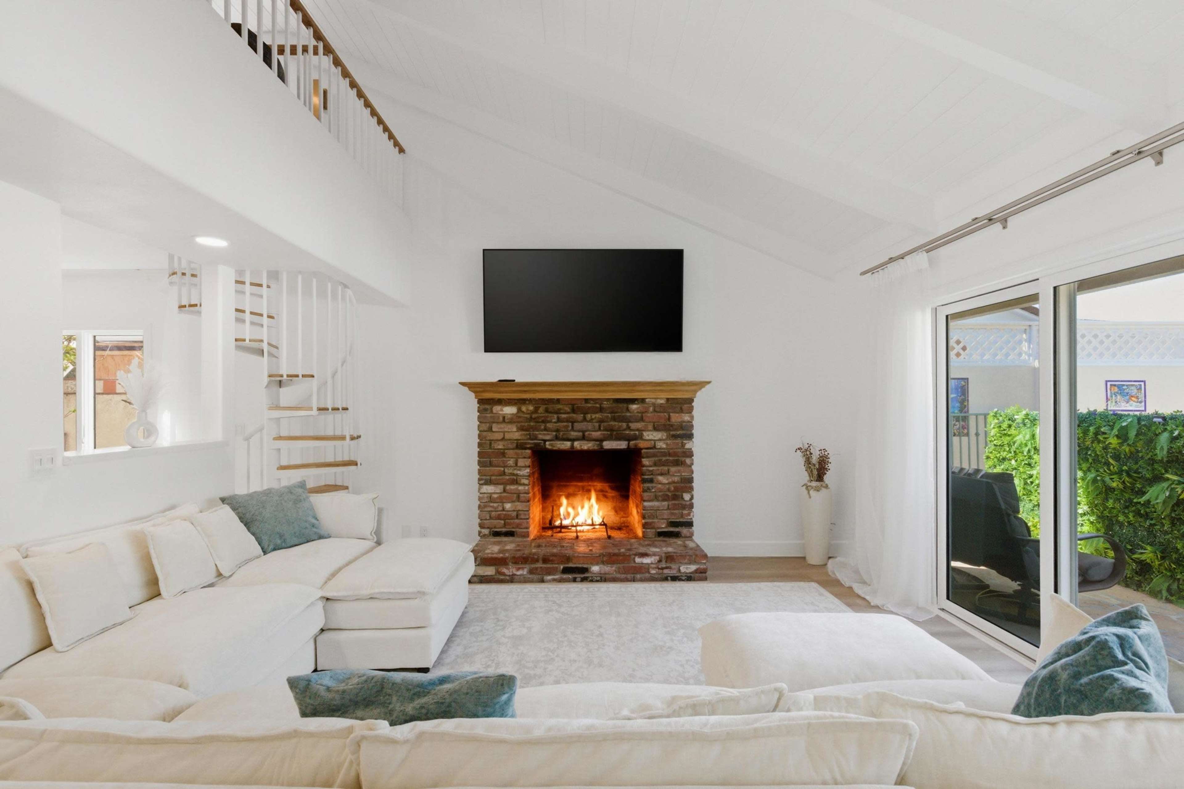 The living room features a white sectional sofa, a brick fireplace with a flame, and a mounted television on a wall with vaulted ceilings.