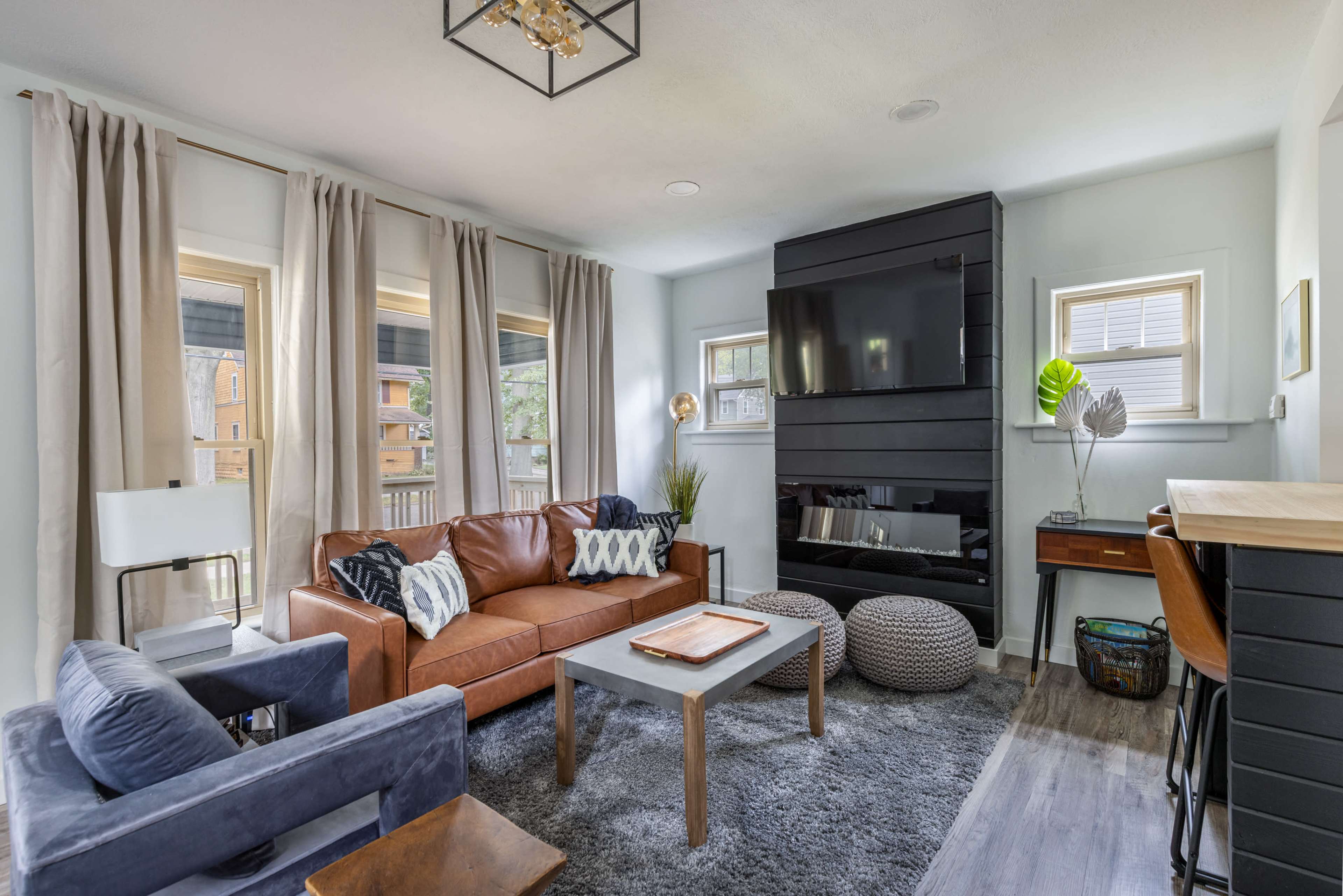 A modern living room features a brown leather sofa, a gray coffee table, a large flat-screen TV mounted above a black fireplace, and floor-to-ceiling curtains.