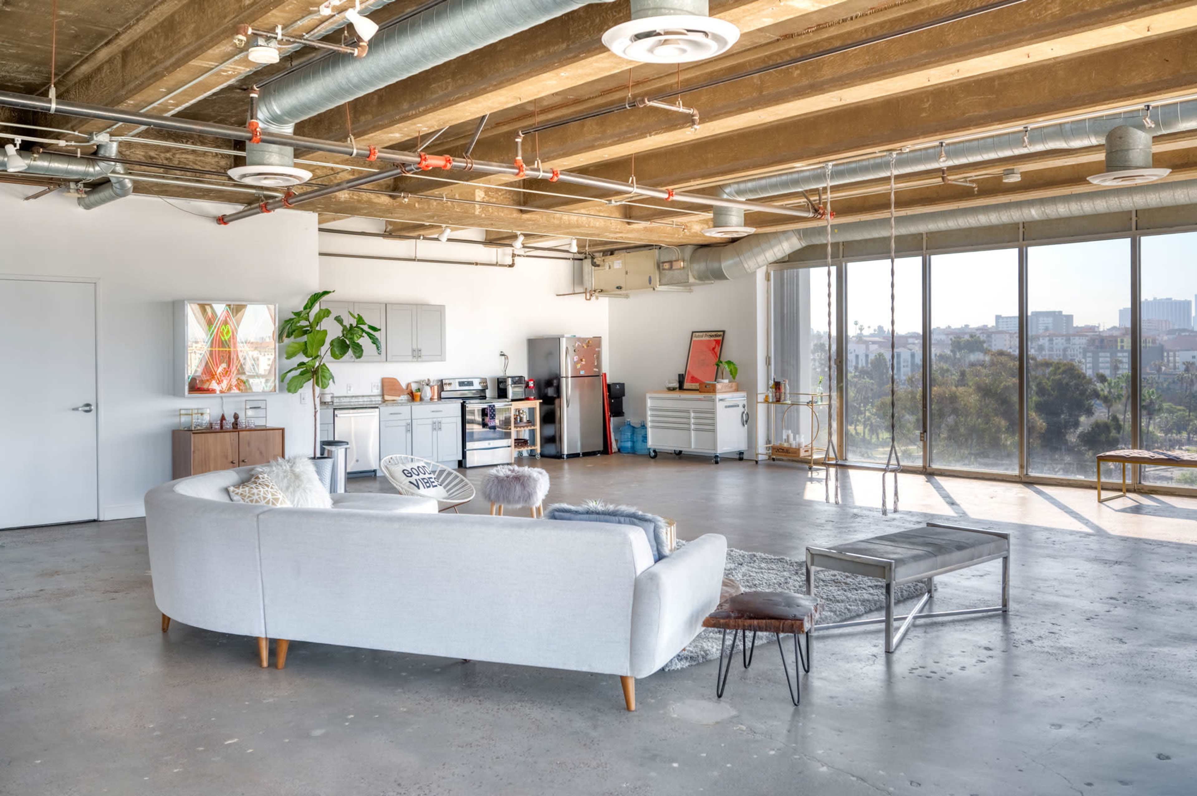 The image shows a spacious, modern living room with industrial-style ceilings, a gray sectional sofa, and large windows overlooking a view of the skyline.