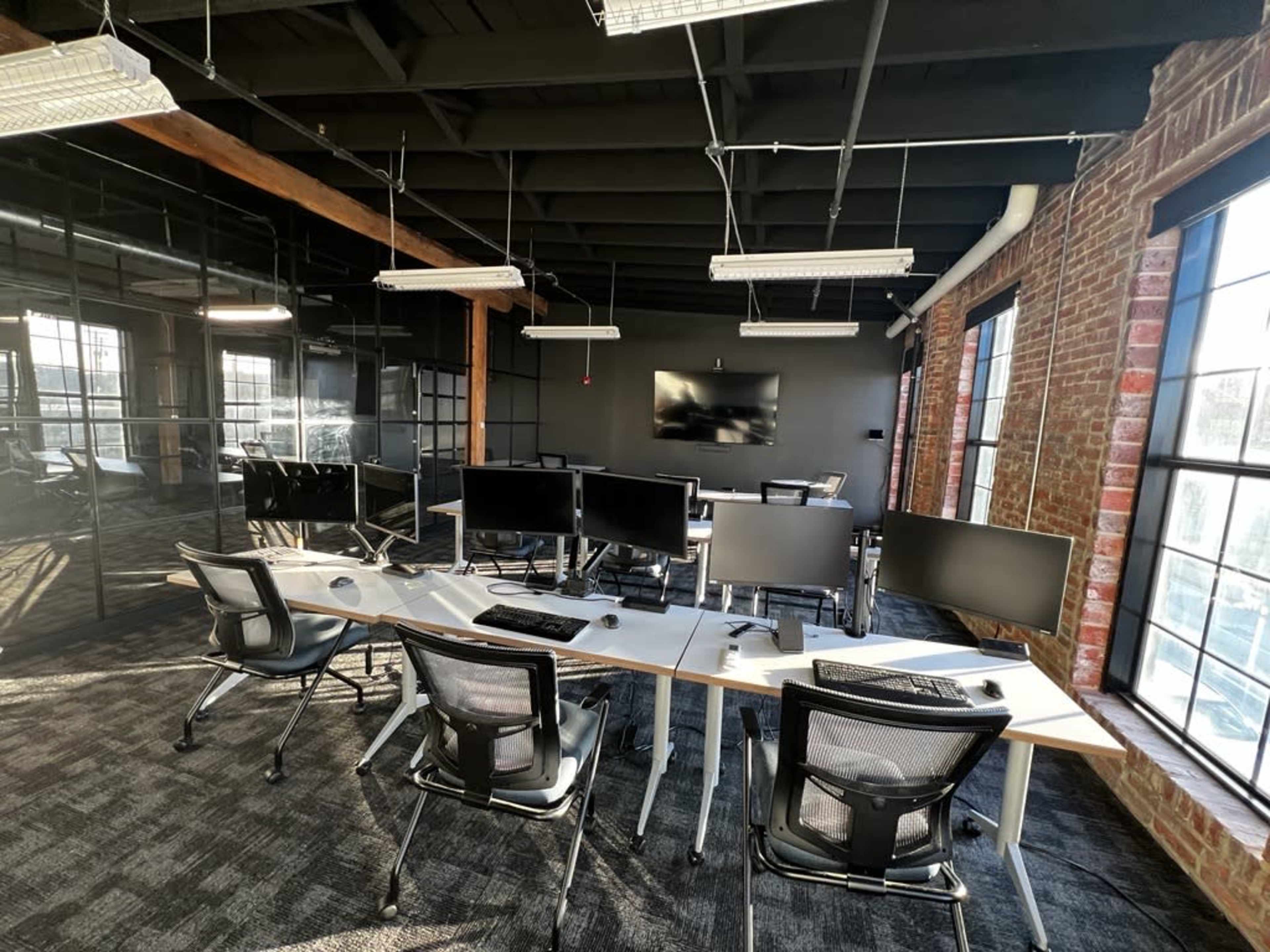 A modern office space with several workstations equipped with desktop monitors, surrounded by large windows and exposed brick walls.