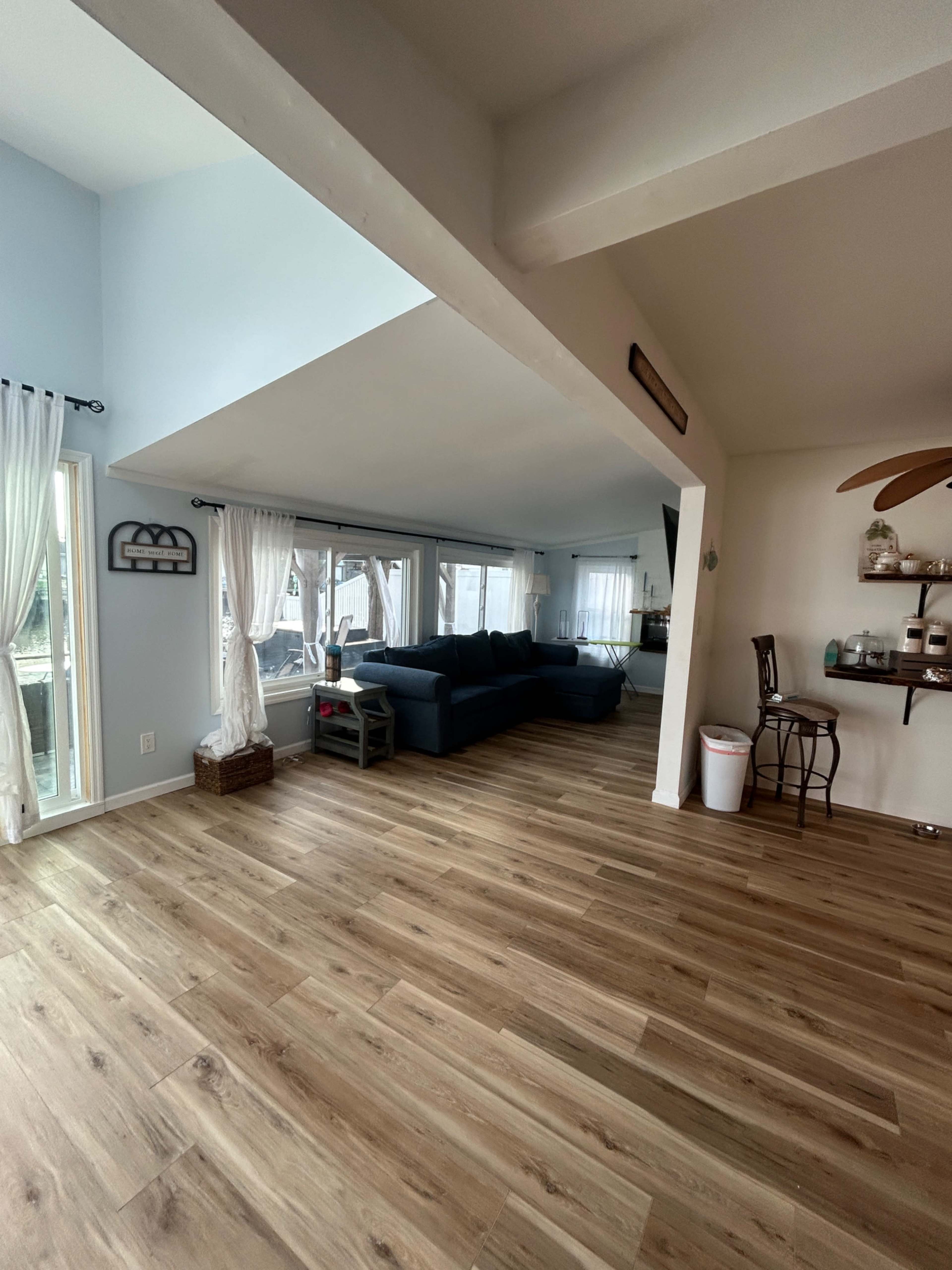 The image shows a spacious living room area with large windows, a dark blue sofa, and wooden flooring.