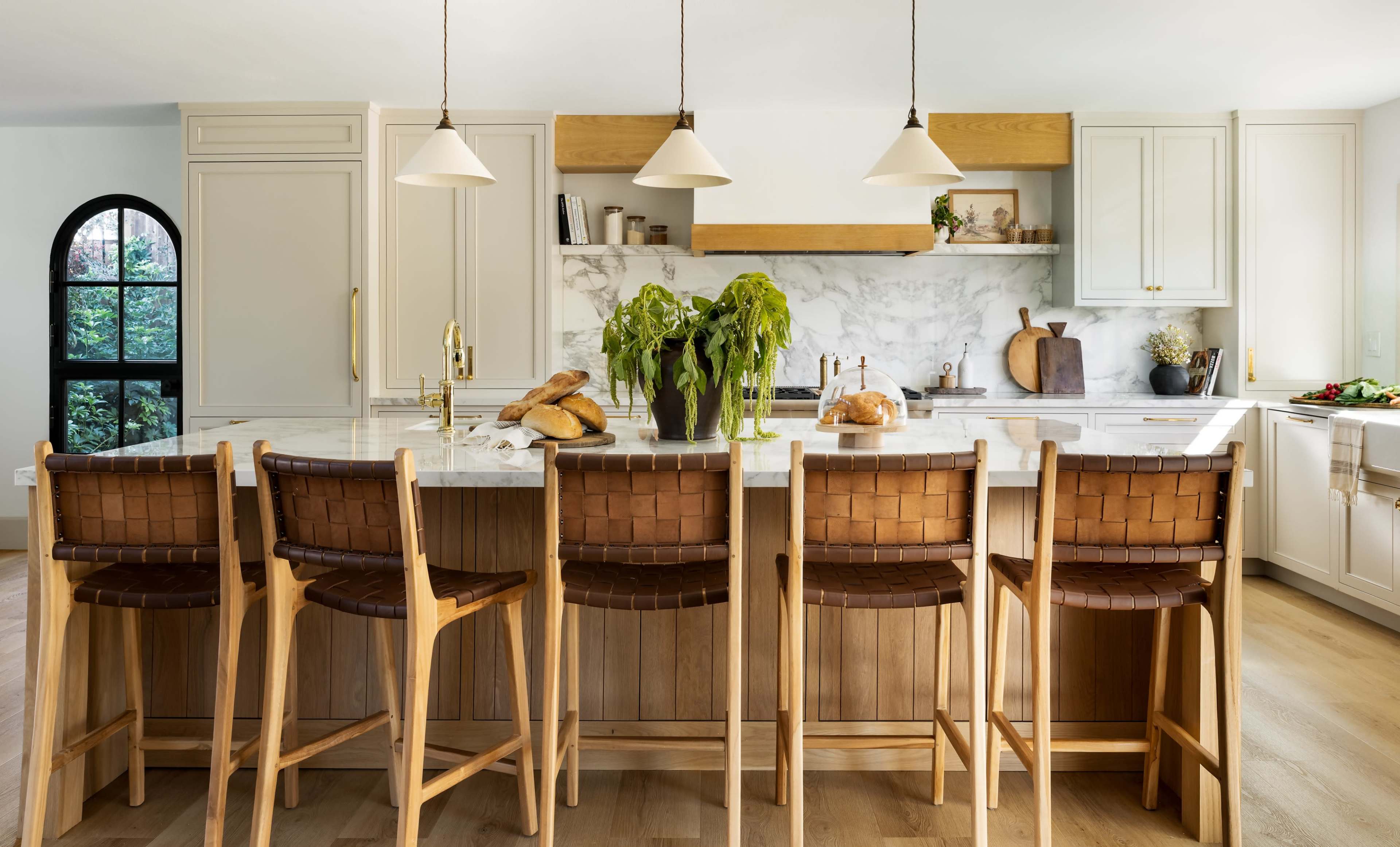A modern kitchen features an island with six bar stools, a marble countertop, and light fixtures hanging above, with plants and baked goods displayed prominently.