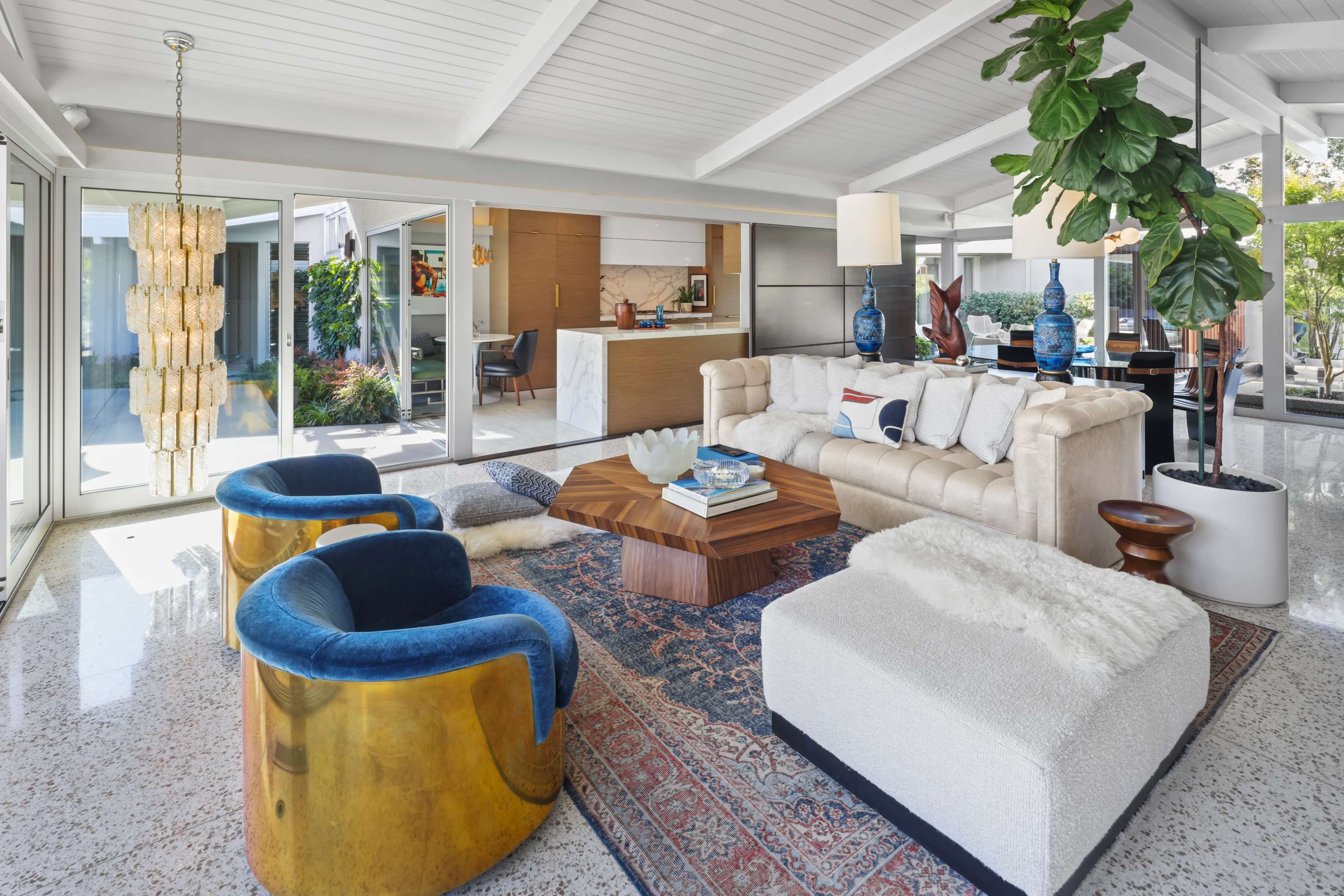 A modern living room with a light-colored sofa, two blue chairs, a wooden coffee table, and large windows that provide natural light.