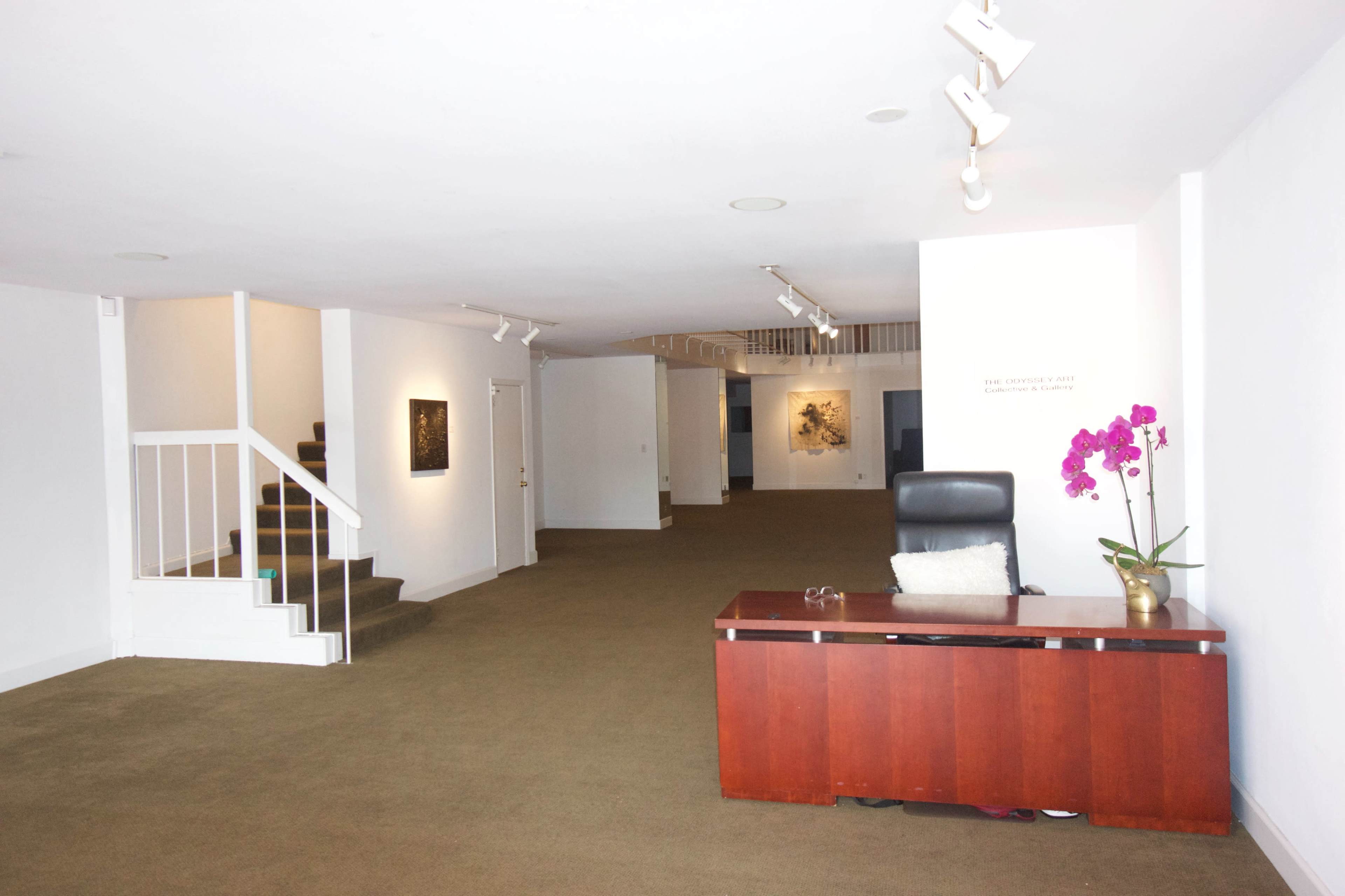The image depicts a spacious, minimalist lobby with a wooden reception desk, a staircase on the left, and artworks displayed on the walls in the background.