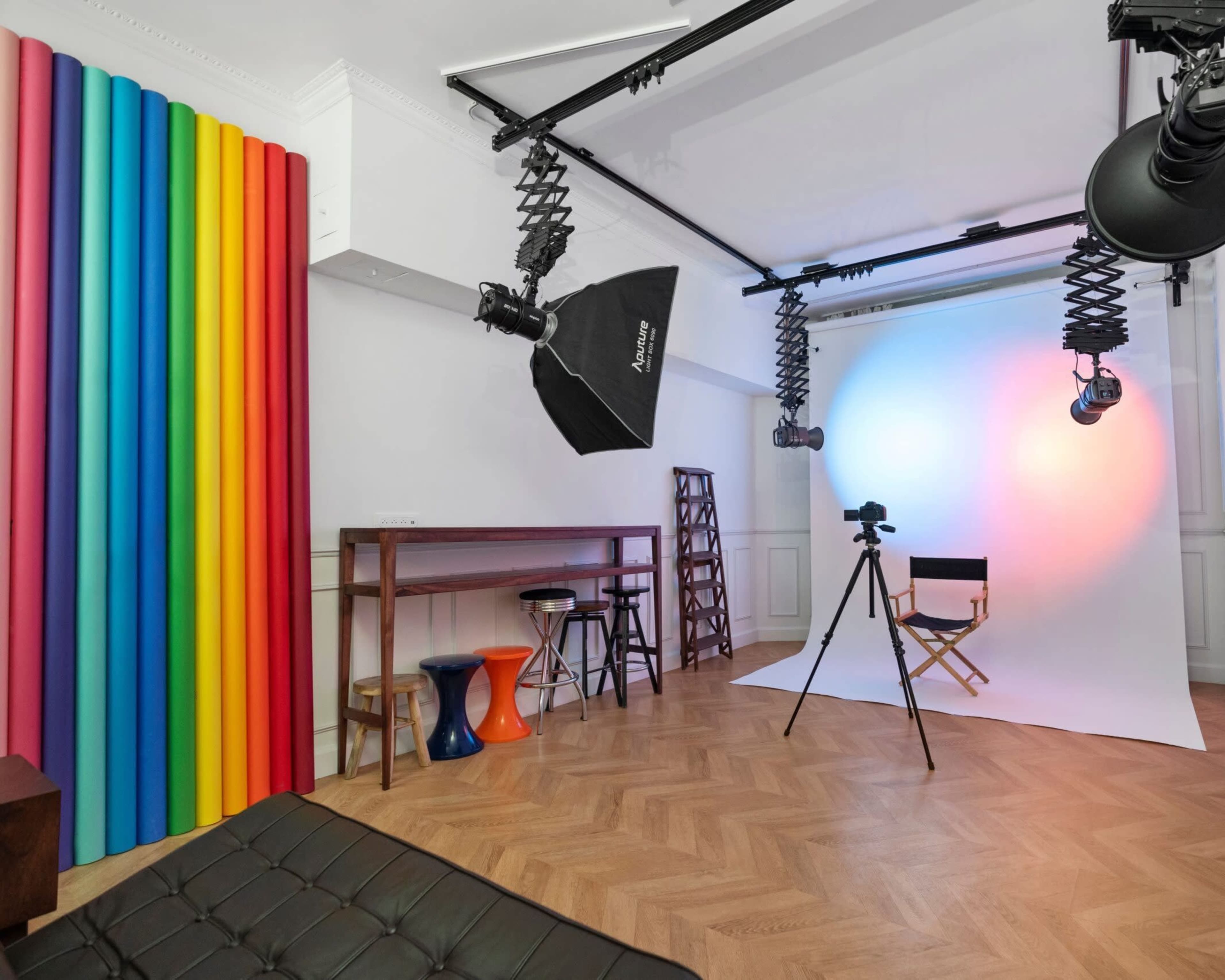 The image shows a photography studio featuring colorful vertical panels, a white backdrop, lighting equipment, and a director's chair.