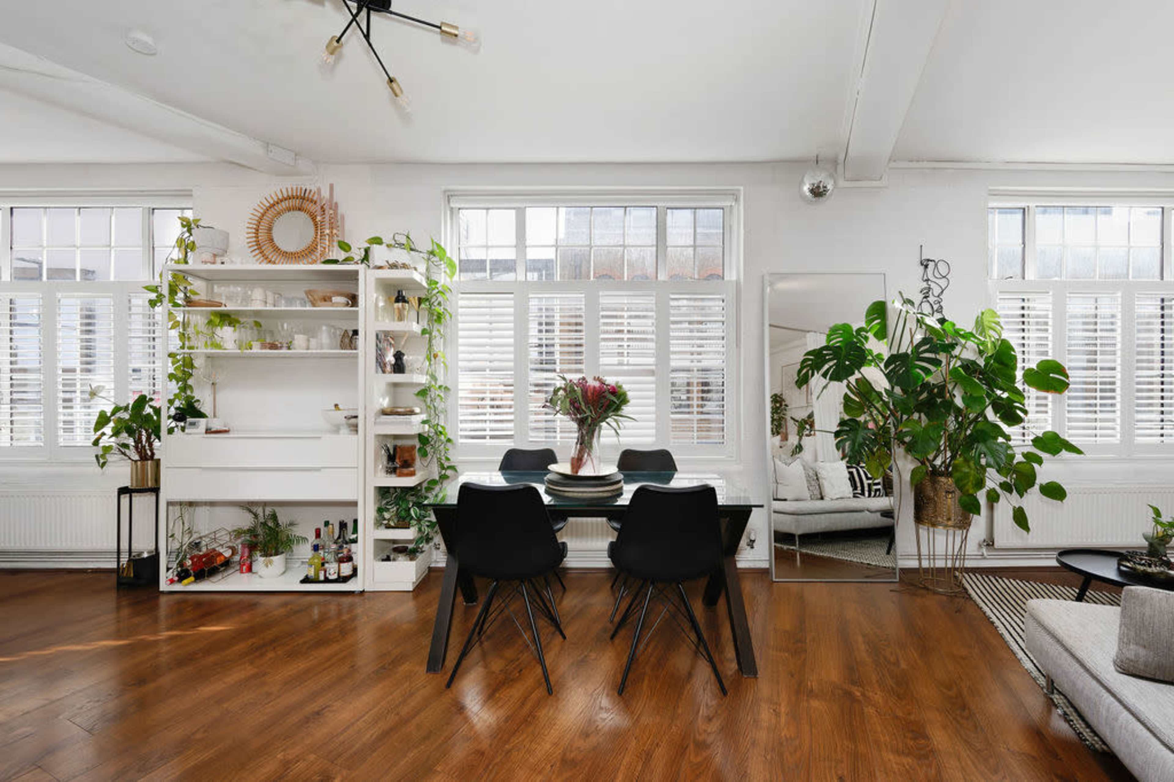 A modern dining area with a black table and chairs, surrounded by plants and shelves against a backdrop of large windows with wooden shutters.