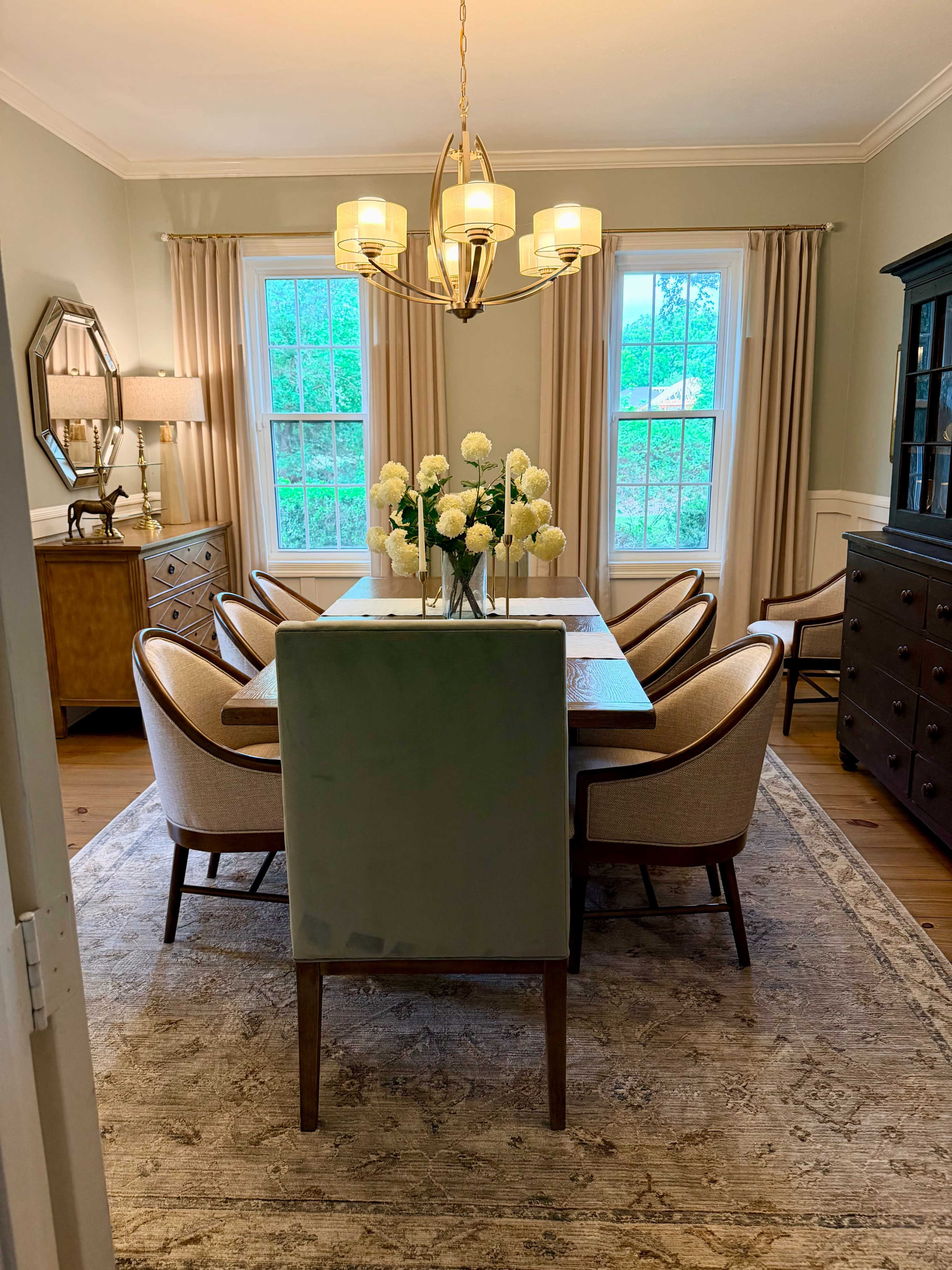A well-furnished dining room features a long table surrounded by upholstered chairs, with large windows allowing natural light and a floral centerpiece.