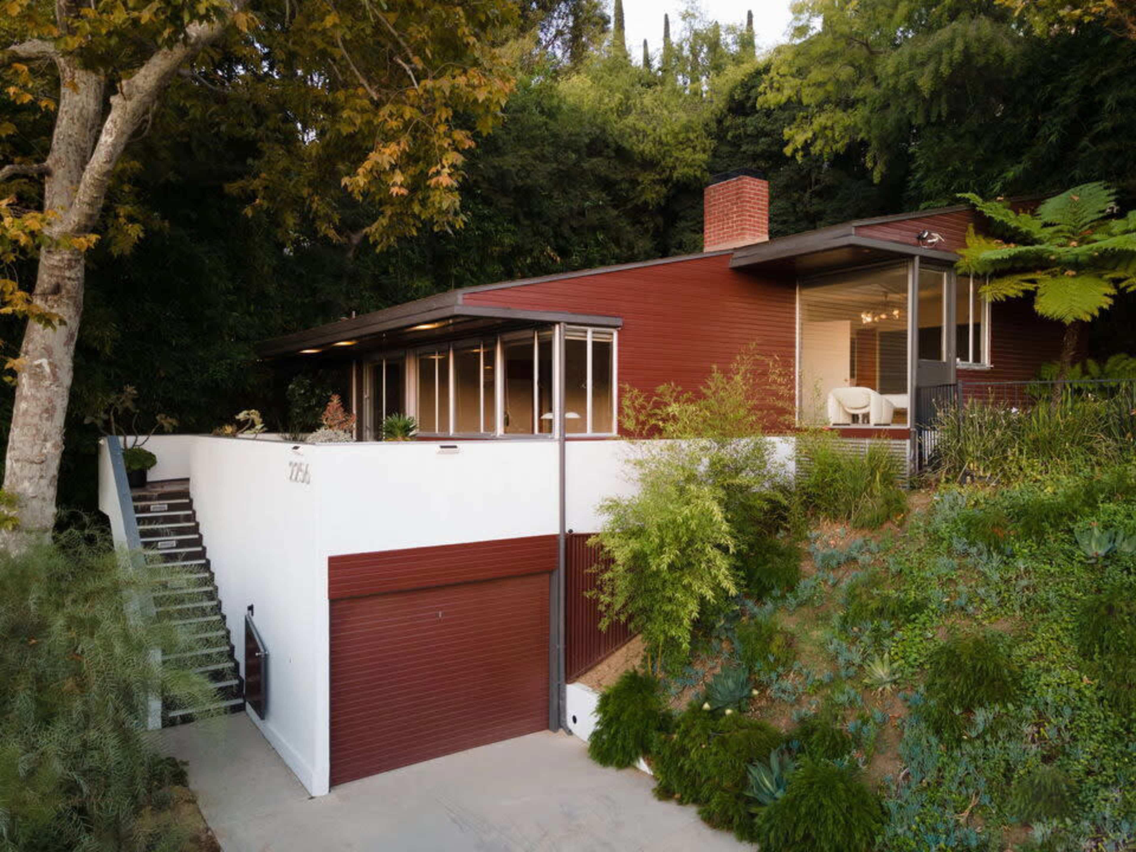A modern red house with large windows is set against a lush hillside, featuring a garage and surrounding greenery.