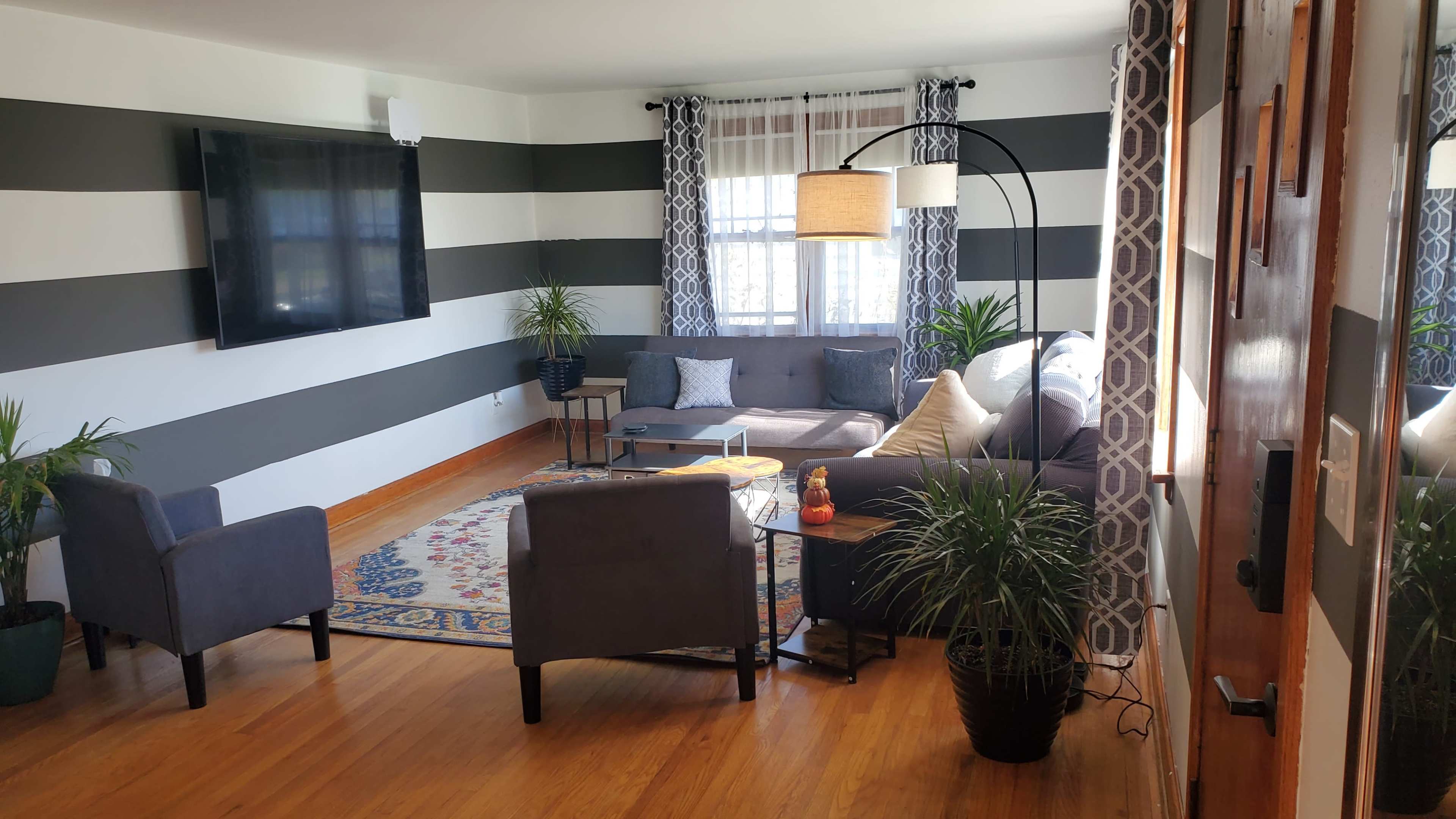 A modern living room with striped walls, a large television, two armchairs, a sofa, and decorative plants.