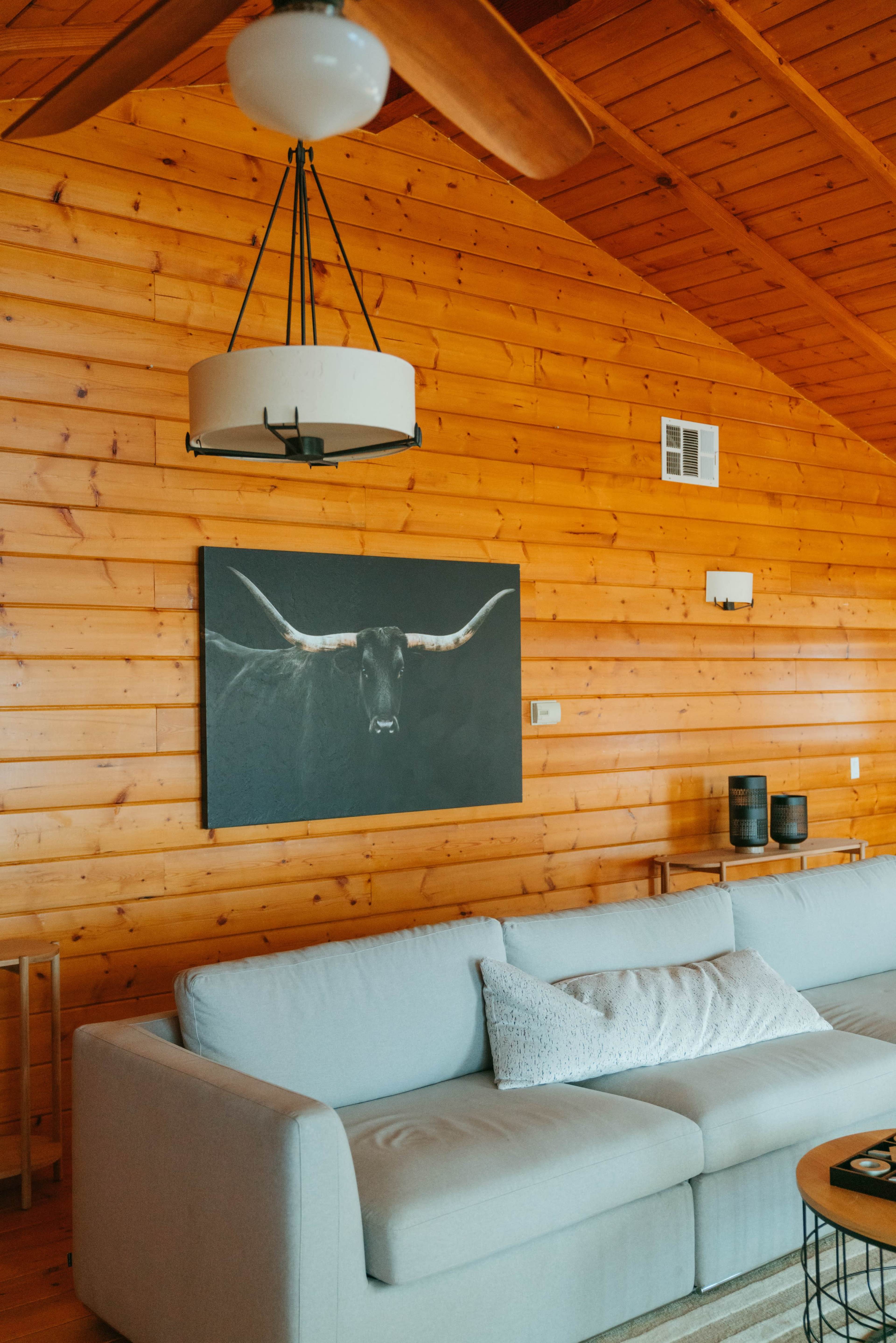 A living room with a light-colored couch, a wooden ceiling, and a large framed artwork of a longhorn bull on a wood-paneled wall.