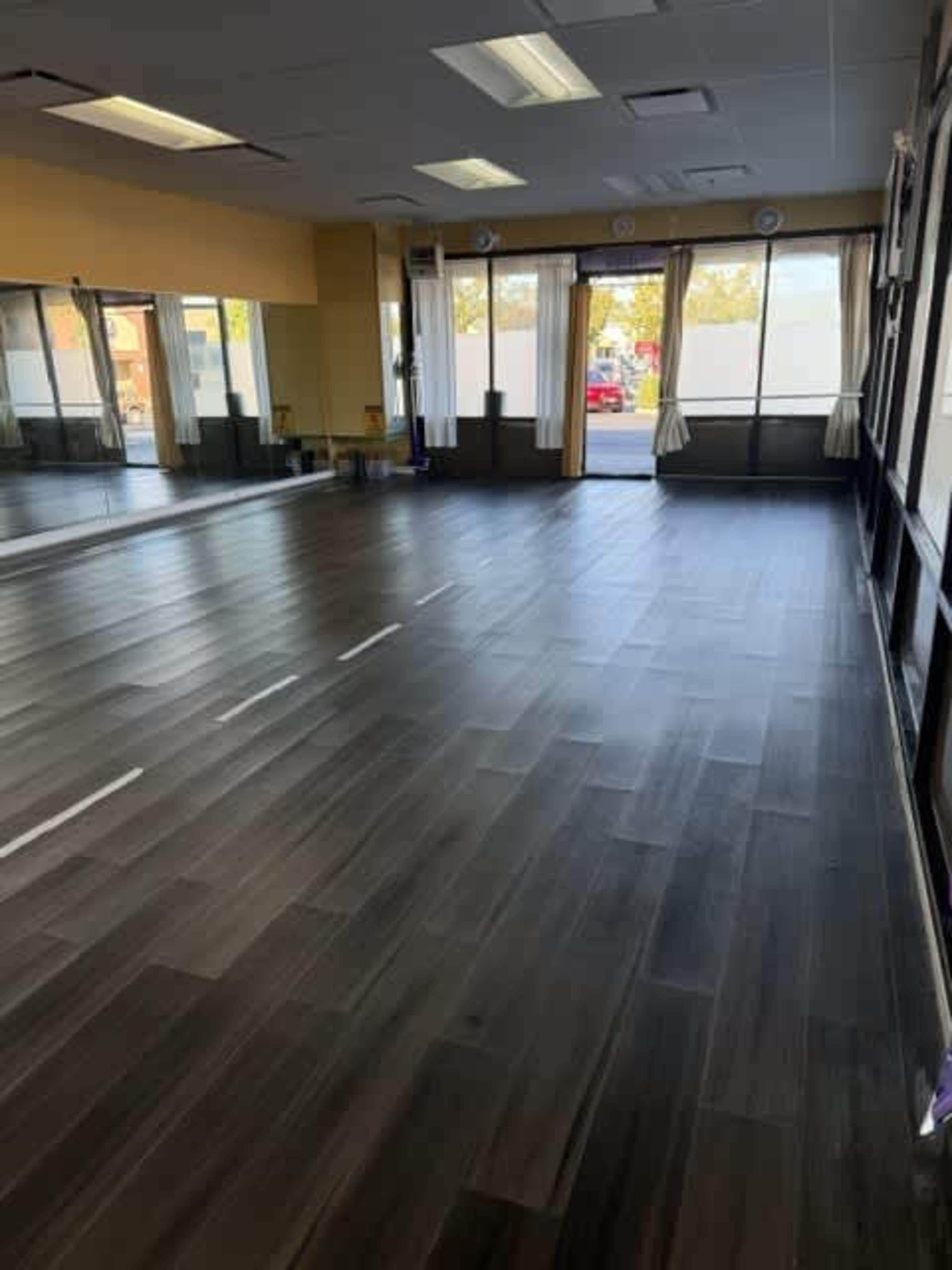 The image shows a spacious studio with wooden flooring, large windows, and mirrors along one wall.