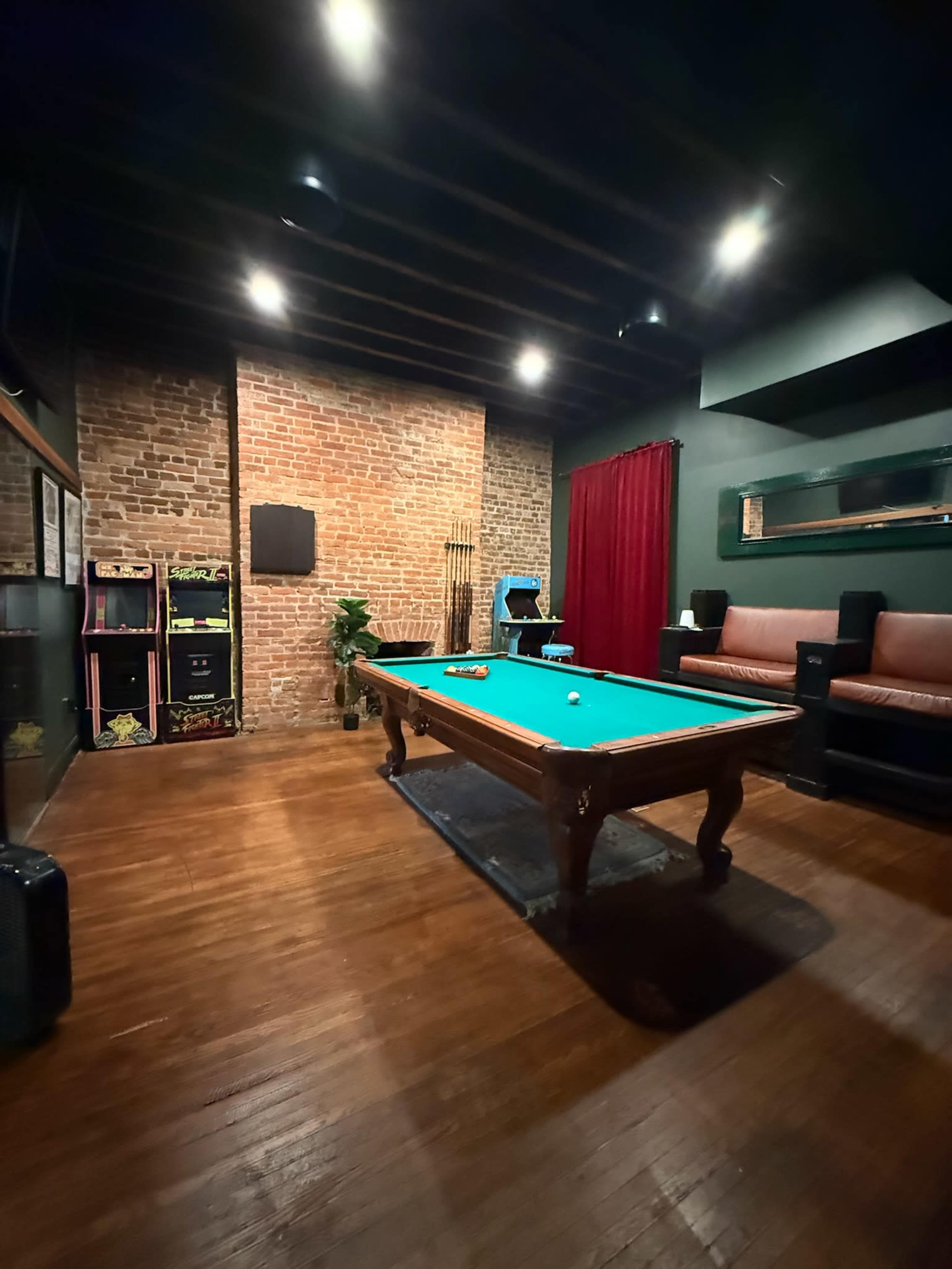 The image shows a game room with a pool table, a brick wall, retro arcade machines, and a seating area with leather couches.