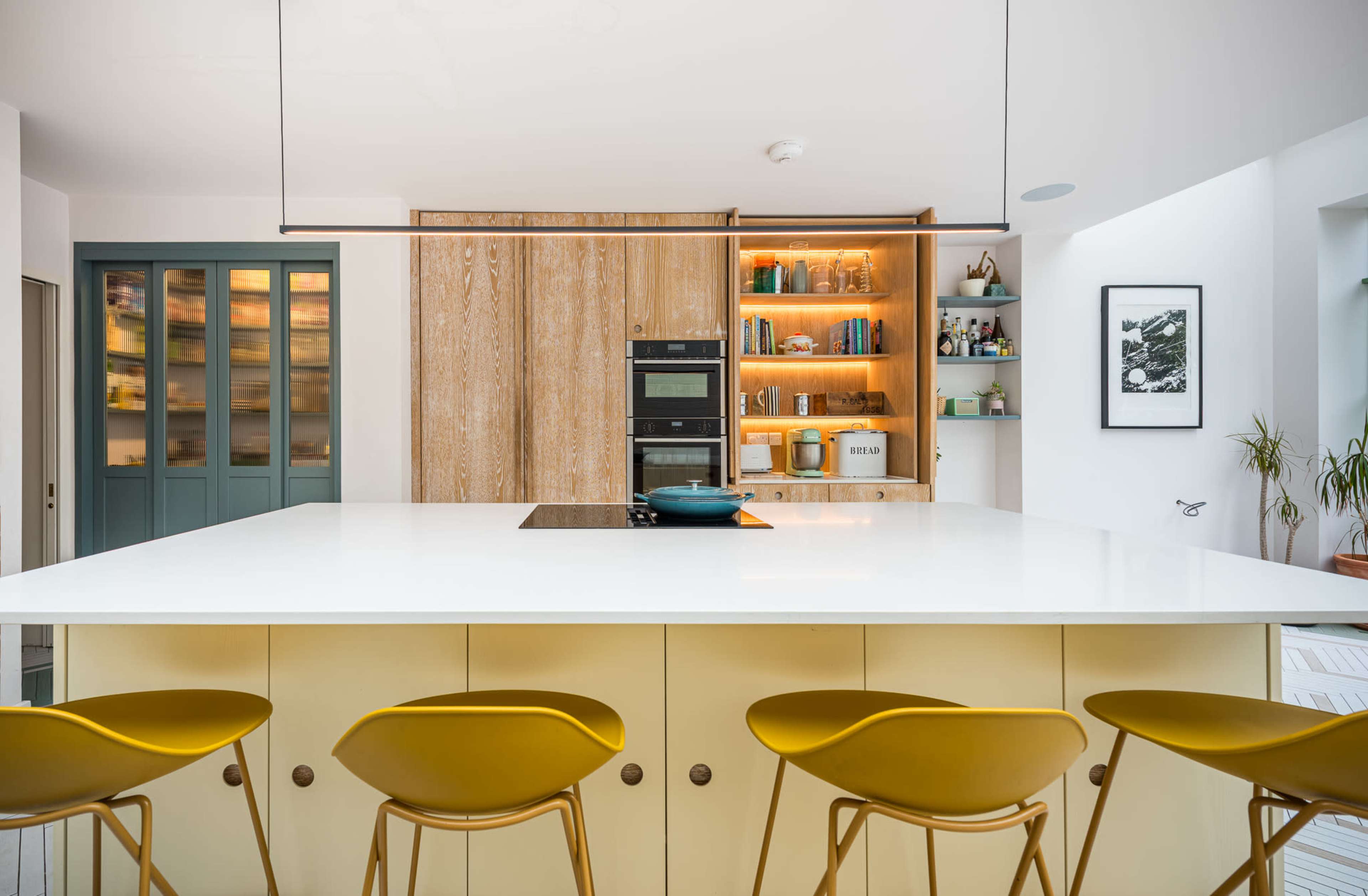 A modern kitchen features a large island with yellow stools, sleek cabinetry, and built-in appliances against a backdrop of warm wood accents.