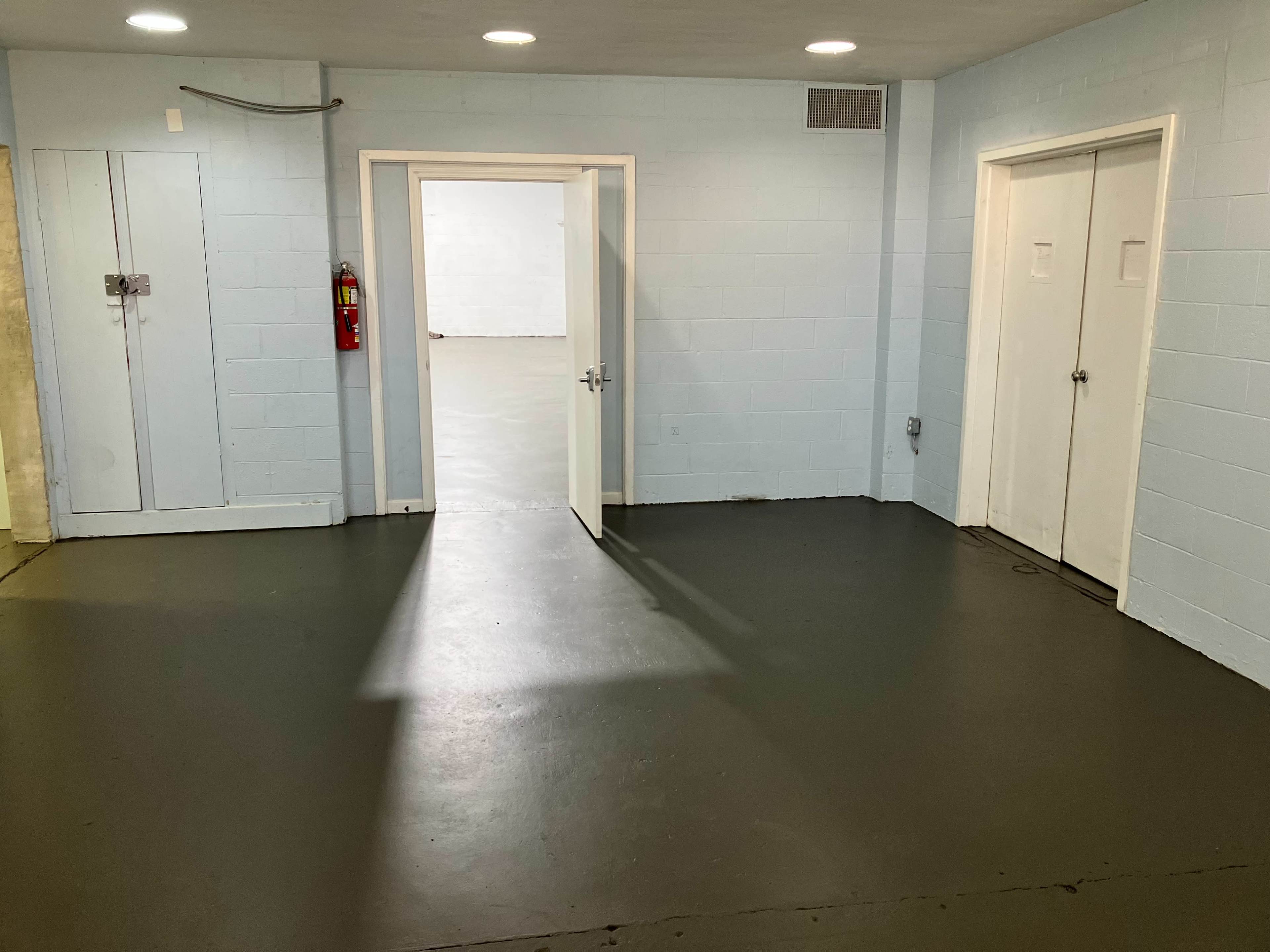 The image shows a plain, empty room with light blue walls, gray flooring, and two closed doors on the right side, along with an open doorway leading to another empty space.