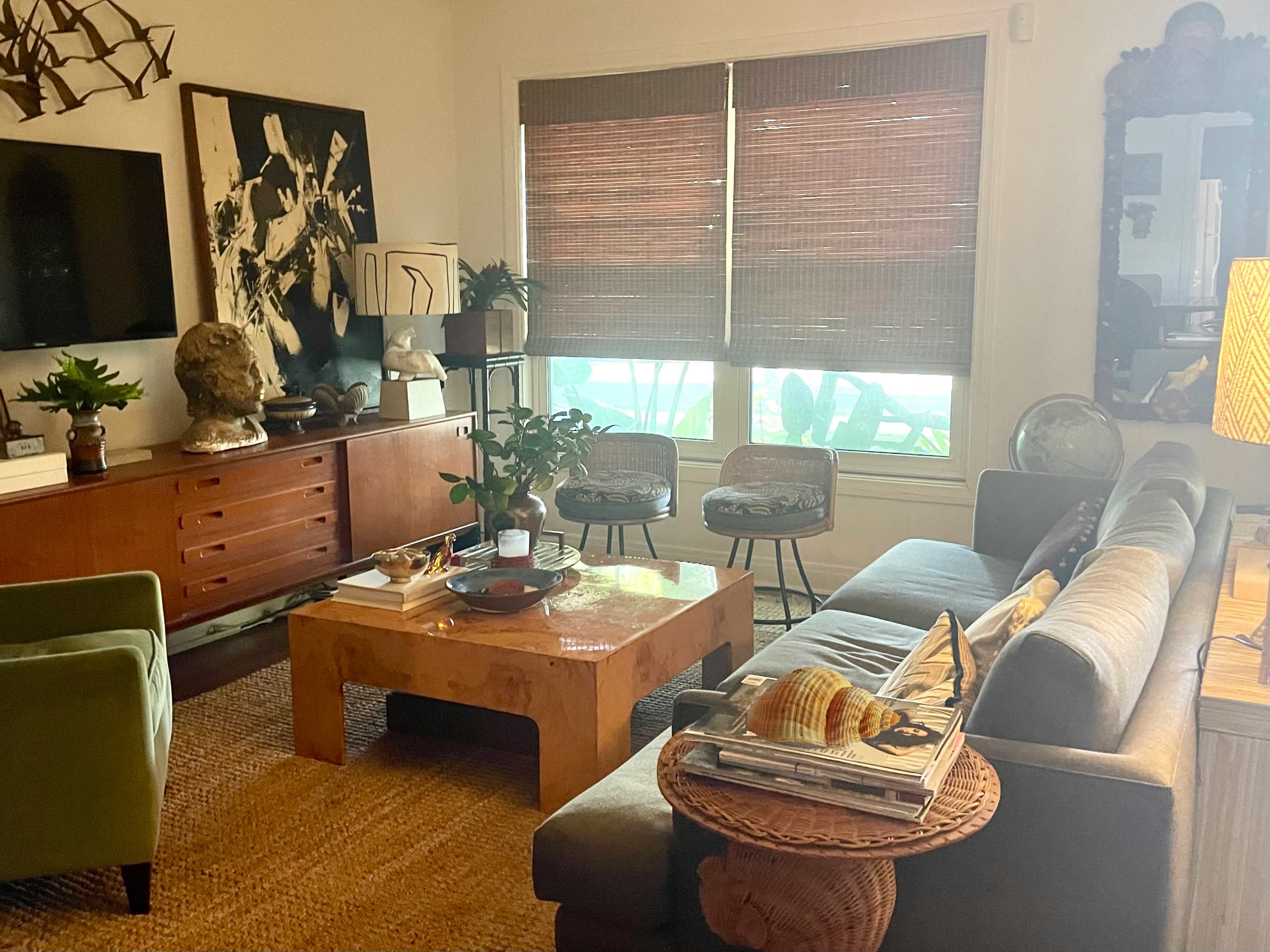 The living room features a sectional sofa, a wooden coffee table, and large windows with bamboo blinds, alongside a mix of decorative plants and artworks.