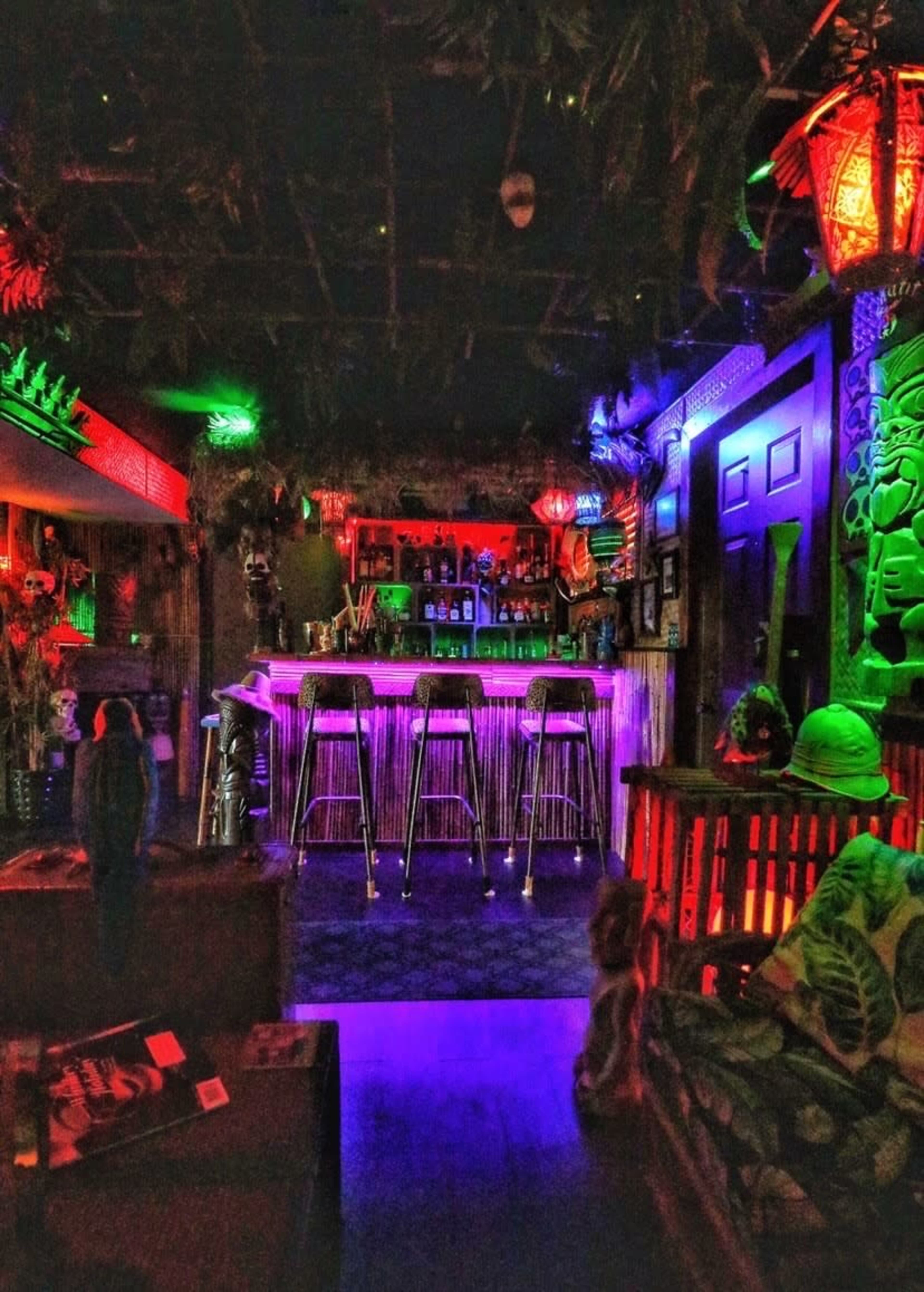 A dimly lit bar area featuring tropical decor, colorful lighting, and high stools arranged around a bar.