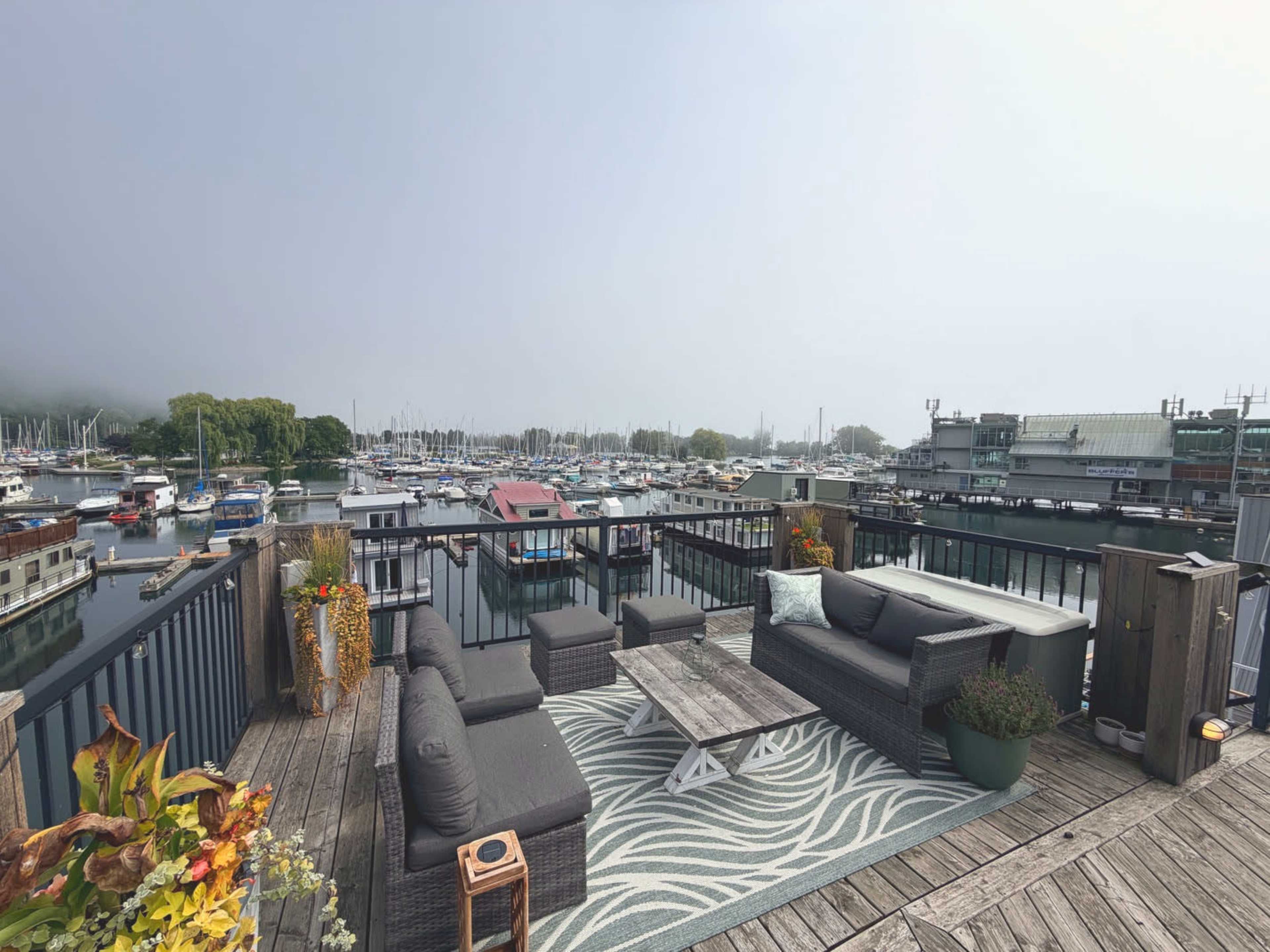 The image shows a balcony area featuring outdoor furniture and a rug, overlooking a marina filled with boats under a cloudy sky.