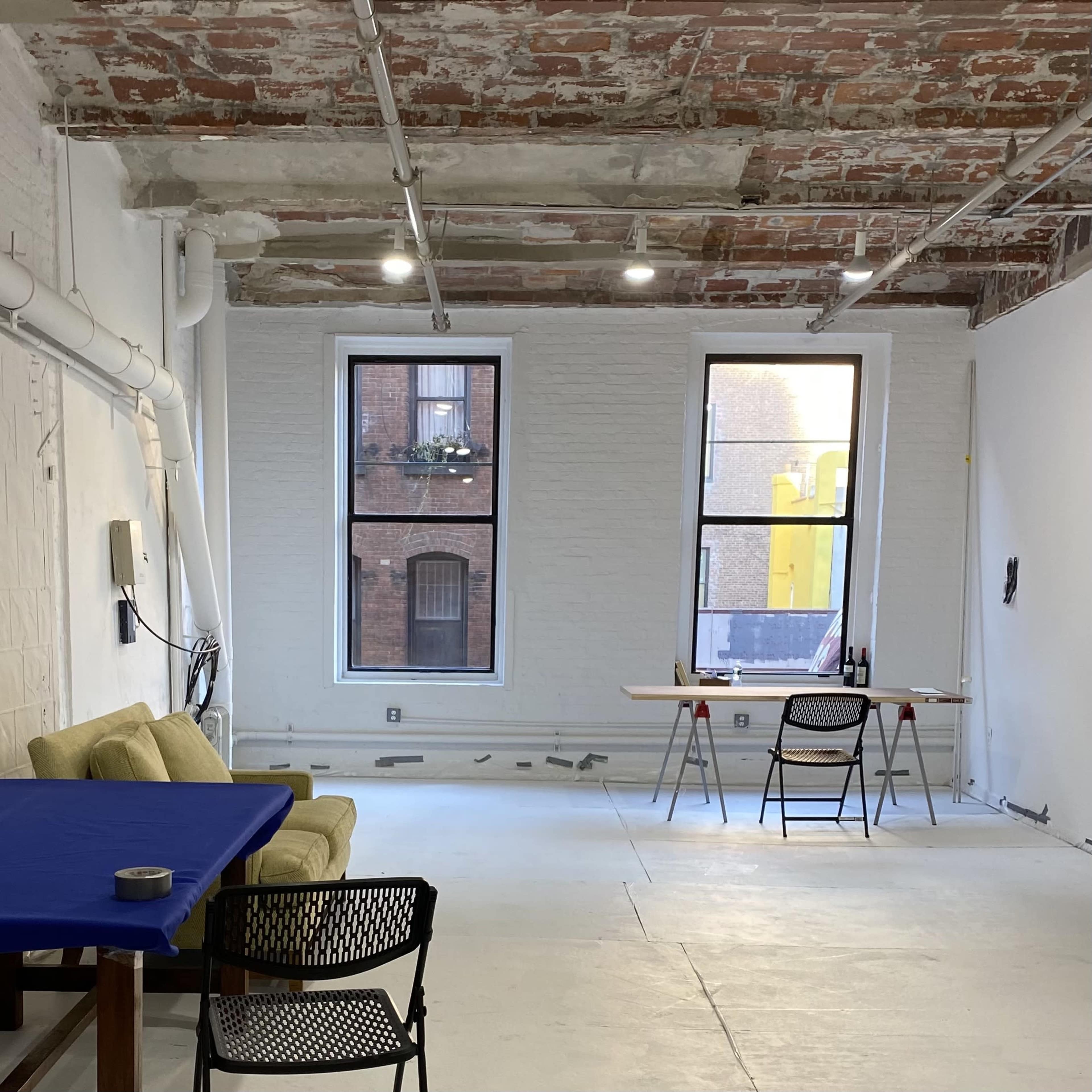 An industrial-style room with exposed brick walls, two large windows, a couch, a table with a chair, and unfinished flooring.