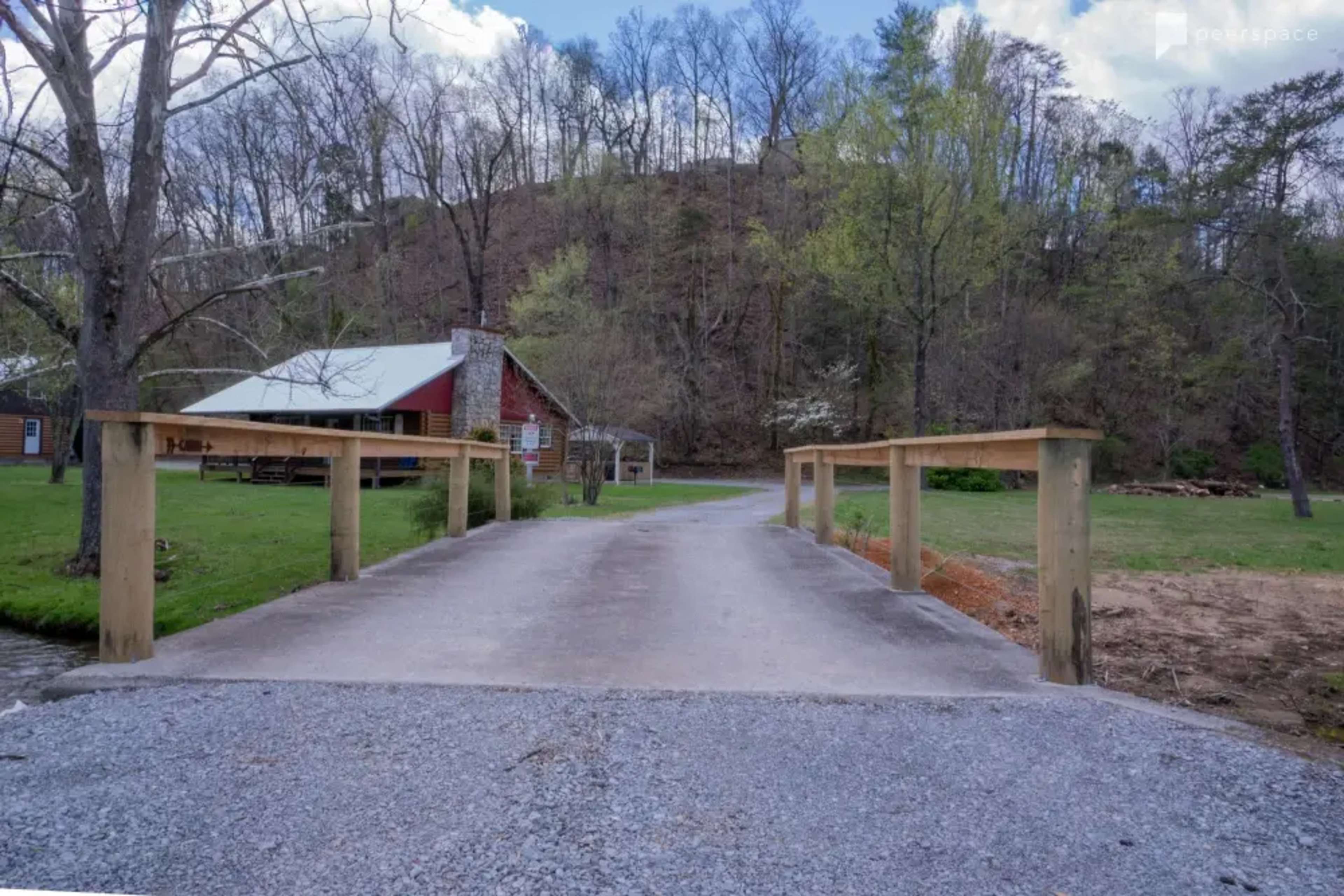 A gravel road leads to a wooden bridge crossing a small stream, with a cabin and trees visible in the background.