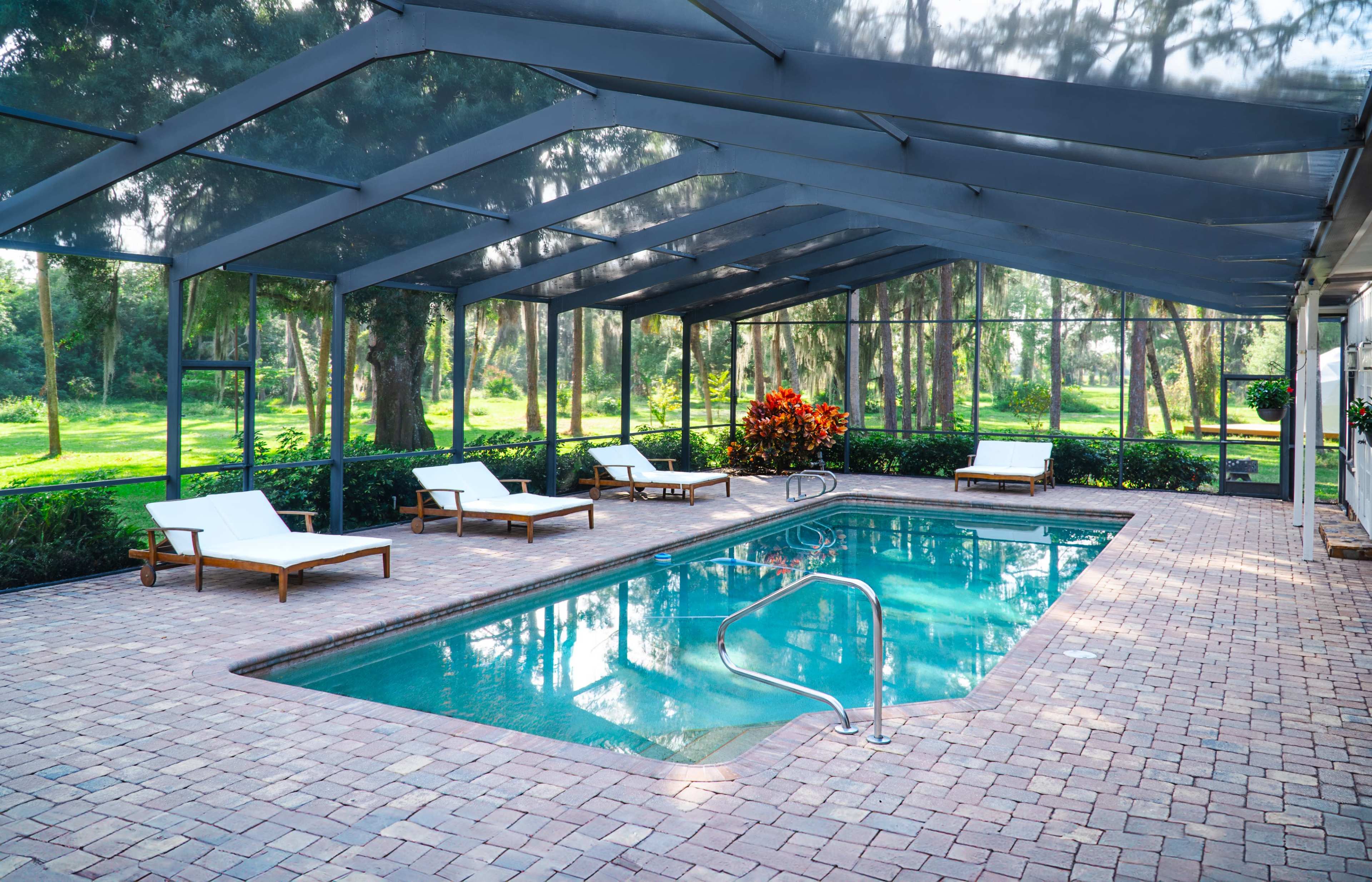 An enclosed pool area with a rectangular swimming pool surrounded by several lounge chairs on a paved surface, set against a backdrop of lush greenery.